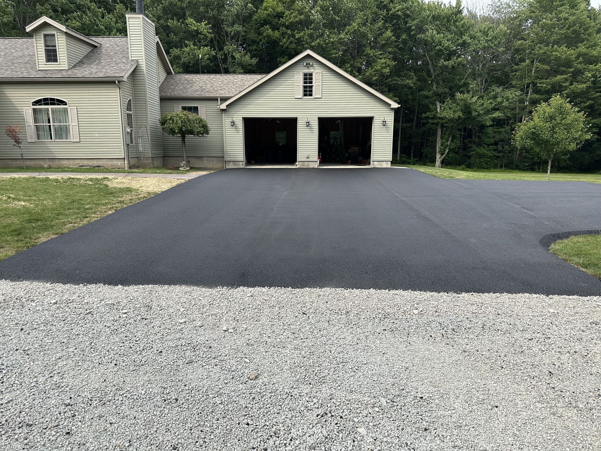 Newly paved asphalt driveway leading to a two-car garage next to a house. Gravel border.