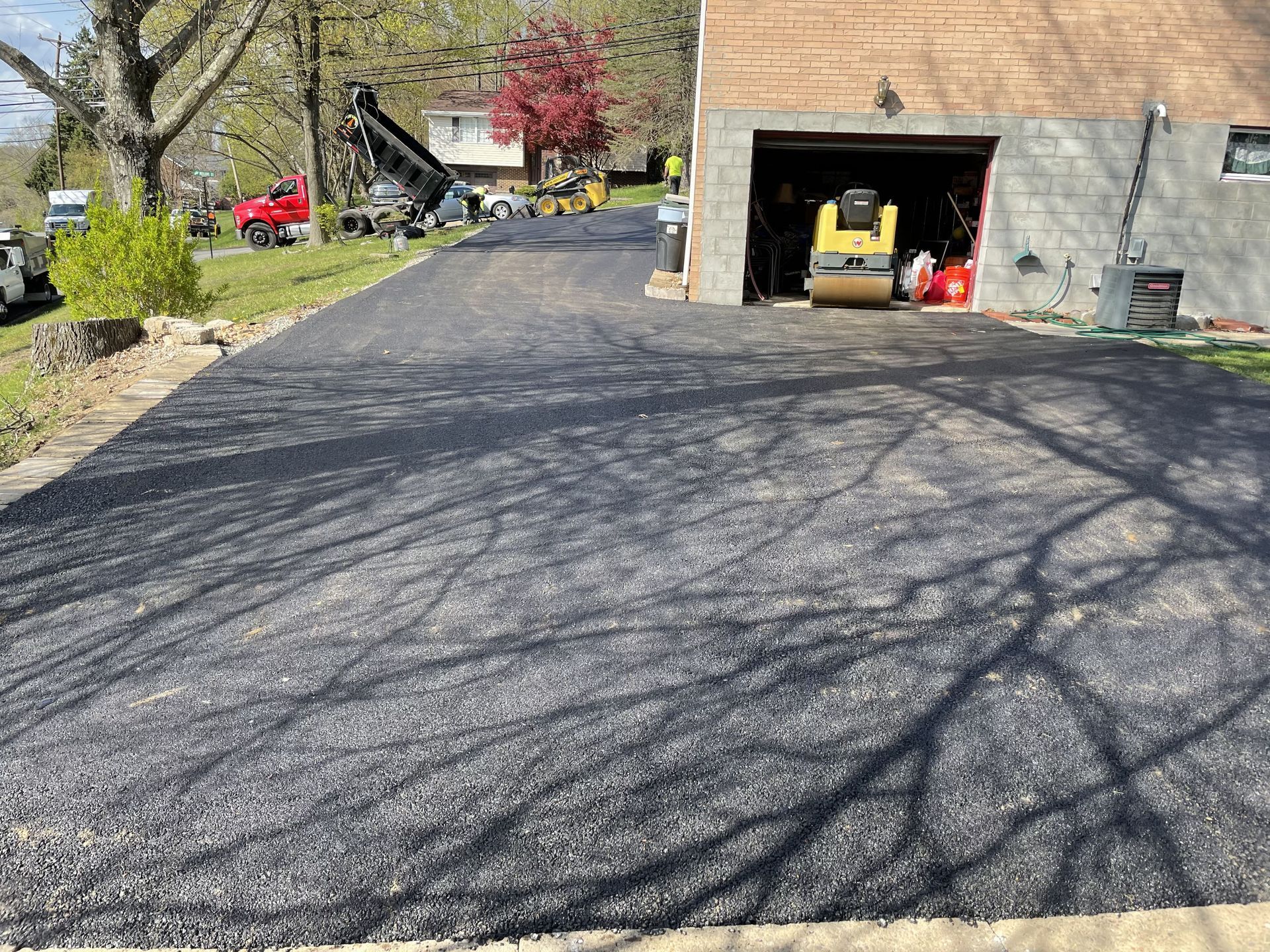 Newly paved asphalt driveway, rolling machine in garage, trees casting shadows.