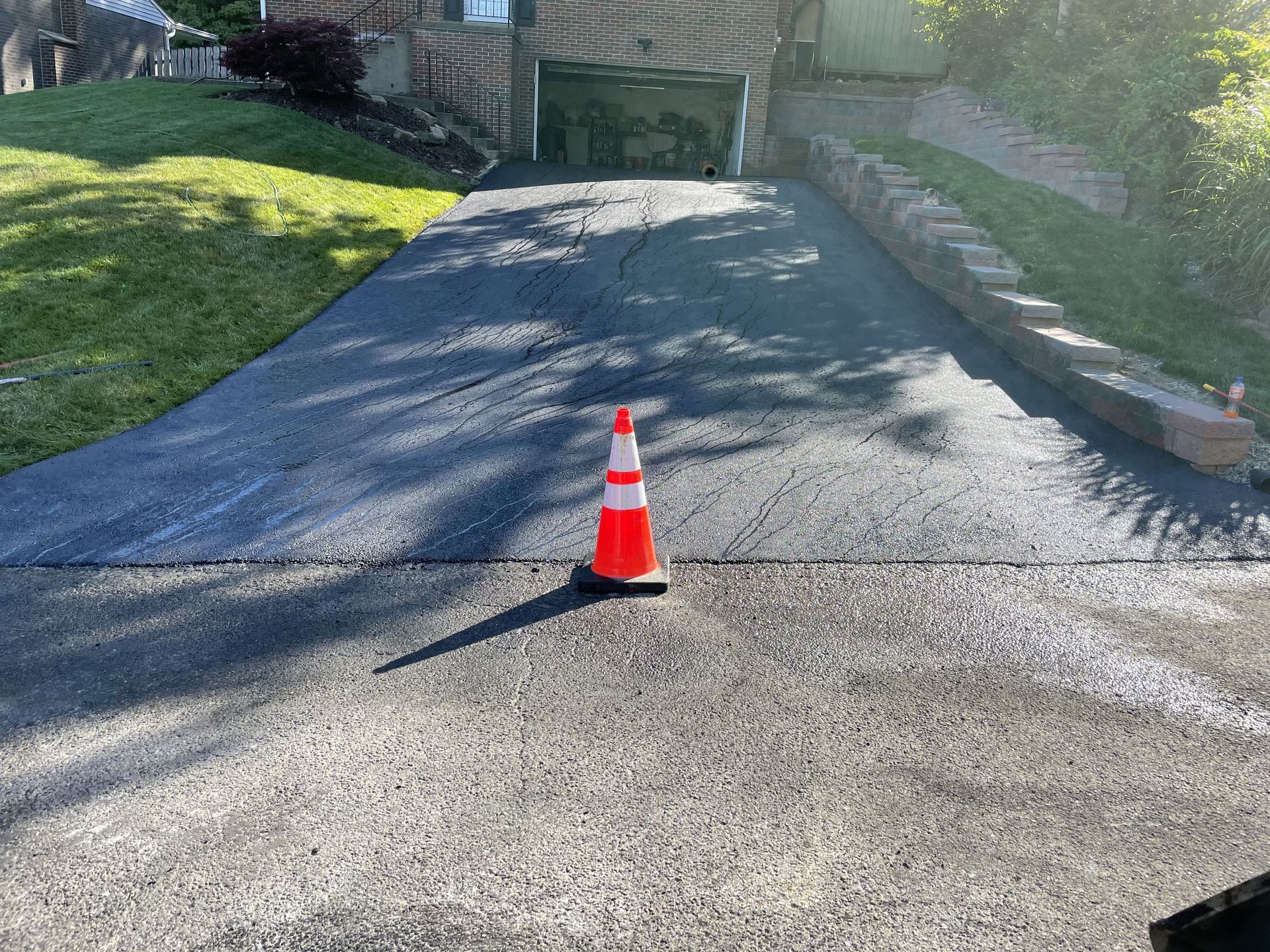 Freshly paved driveway with orange traffic cone in the center. Green grass and stone retaining wall on either side.