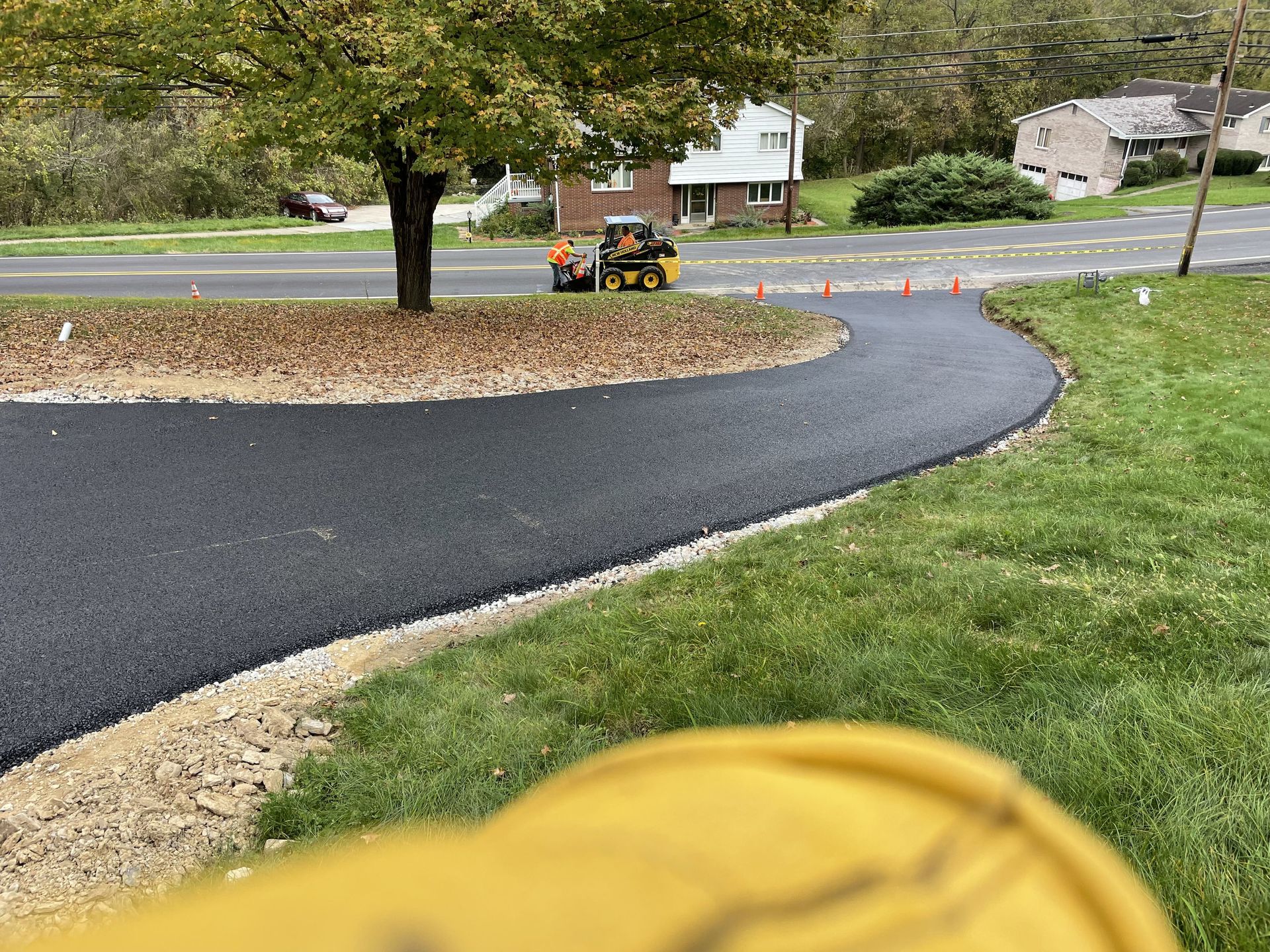 Newly paved black asphalt road with curved edges, surrounded by grass and gravel. A construction vehicle is present.
