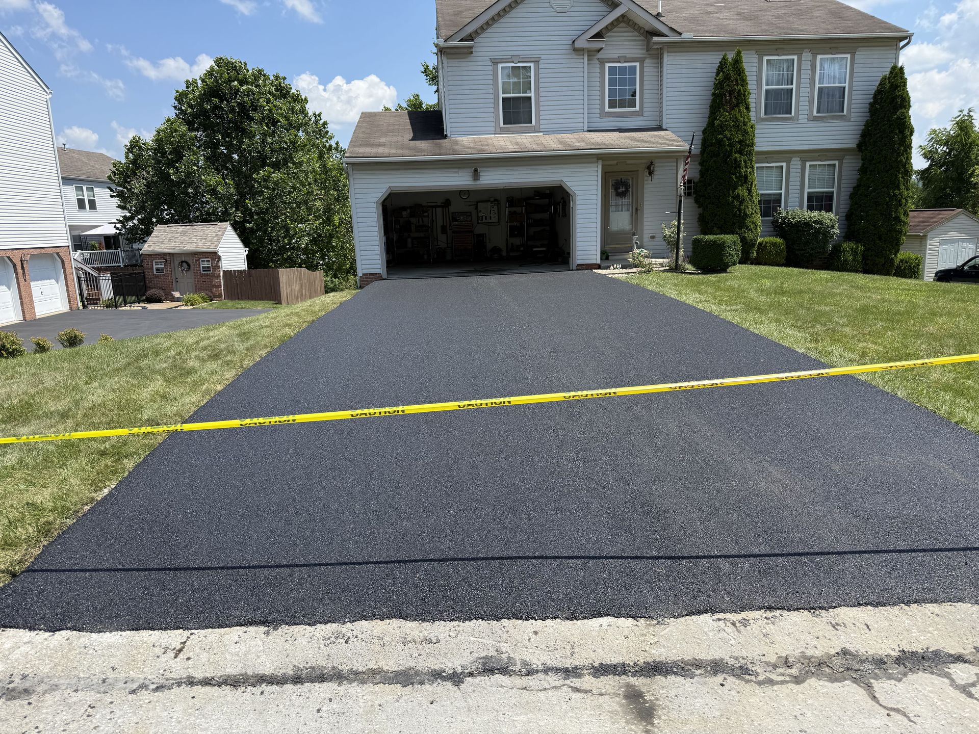 Newly paved black asphalt driveway in front of a two-story house, bordered by grass and caution tape.