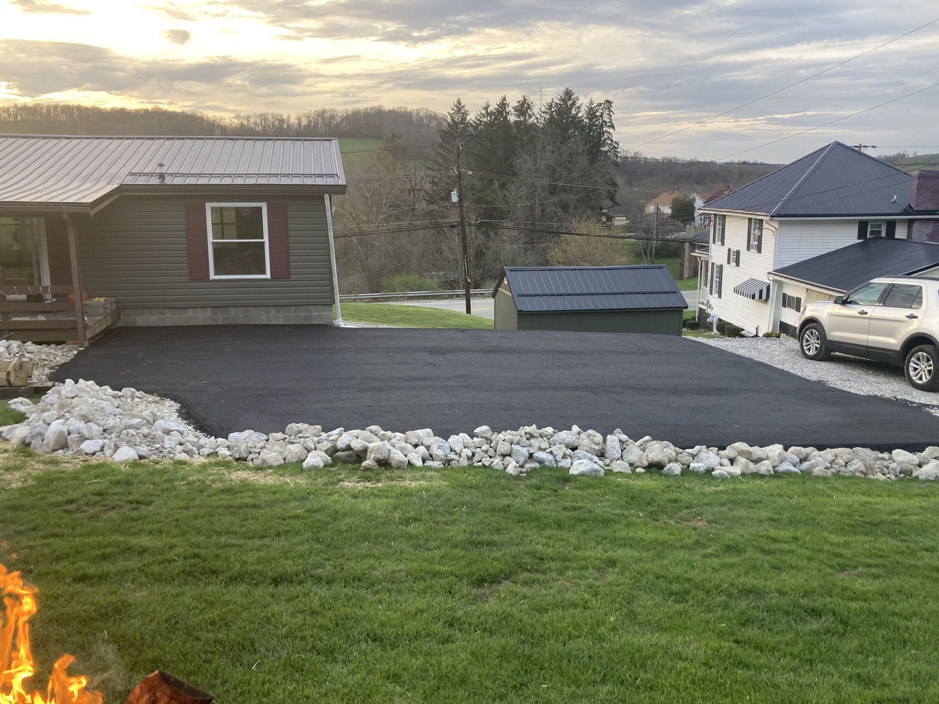Newly paved asphalt driveway with rock border in front of a house, set in a rural landscape.