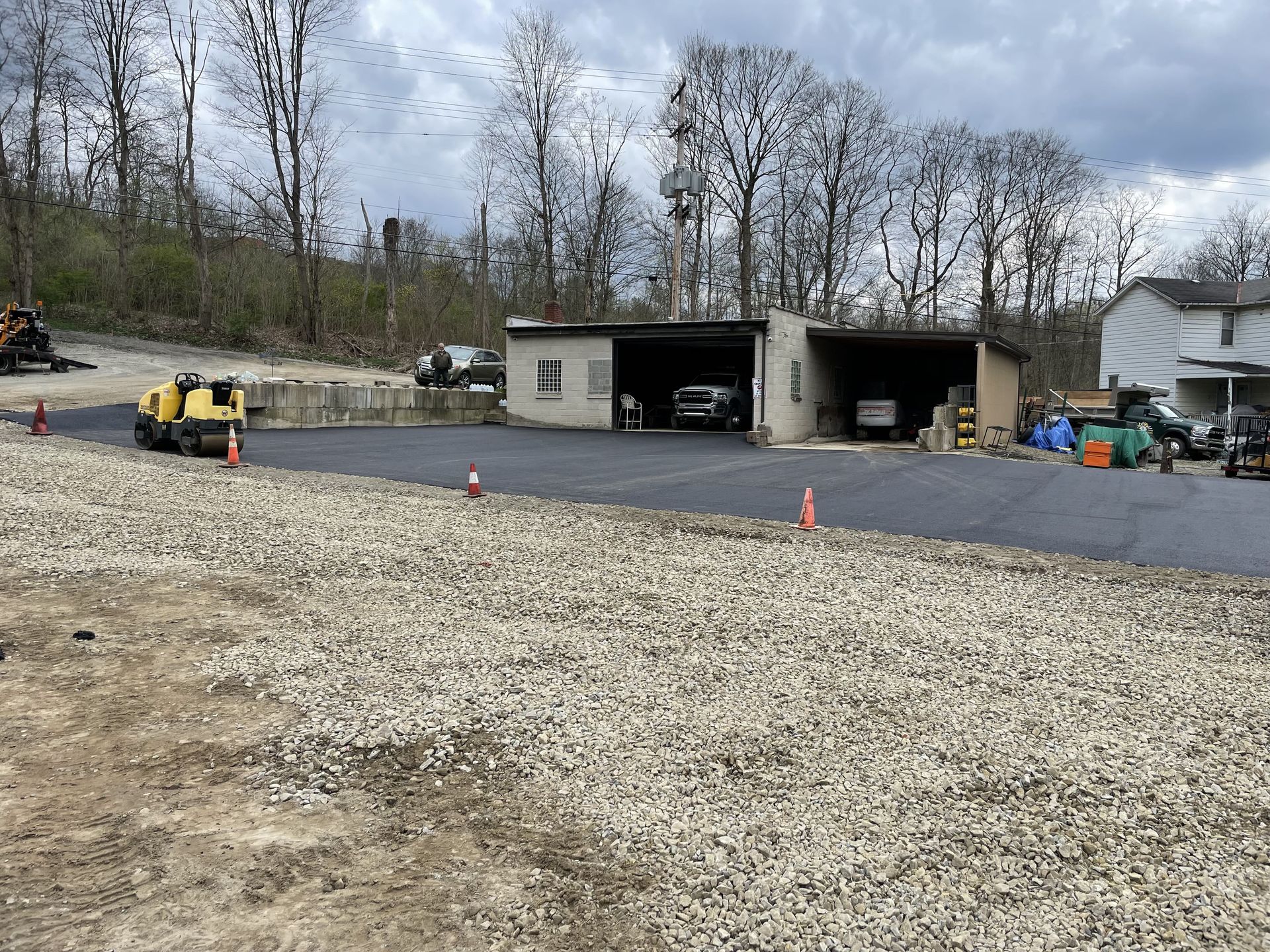 Gravel lot in front of asphalt driveway and garage, construction equipment visible, trees in background.