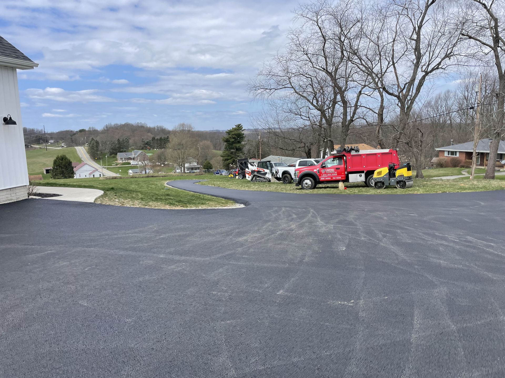New asphalt driveway with parked trucks, grass, and houses in the background under a cloudy sky.