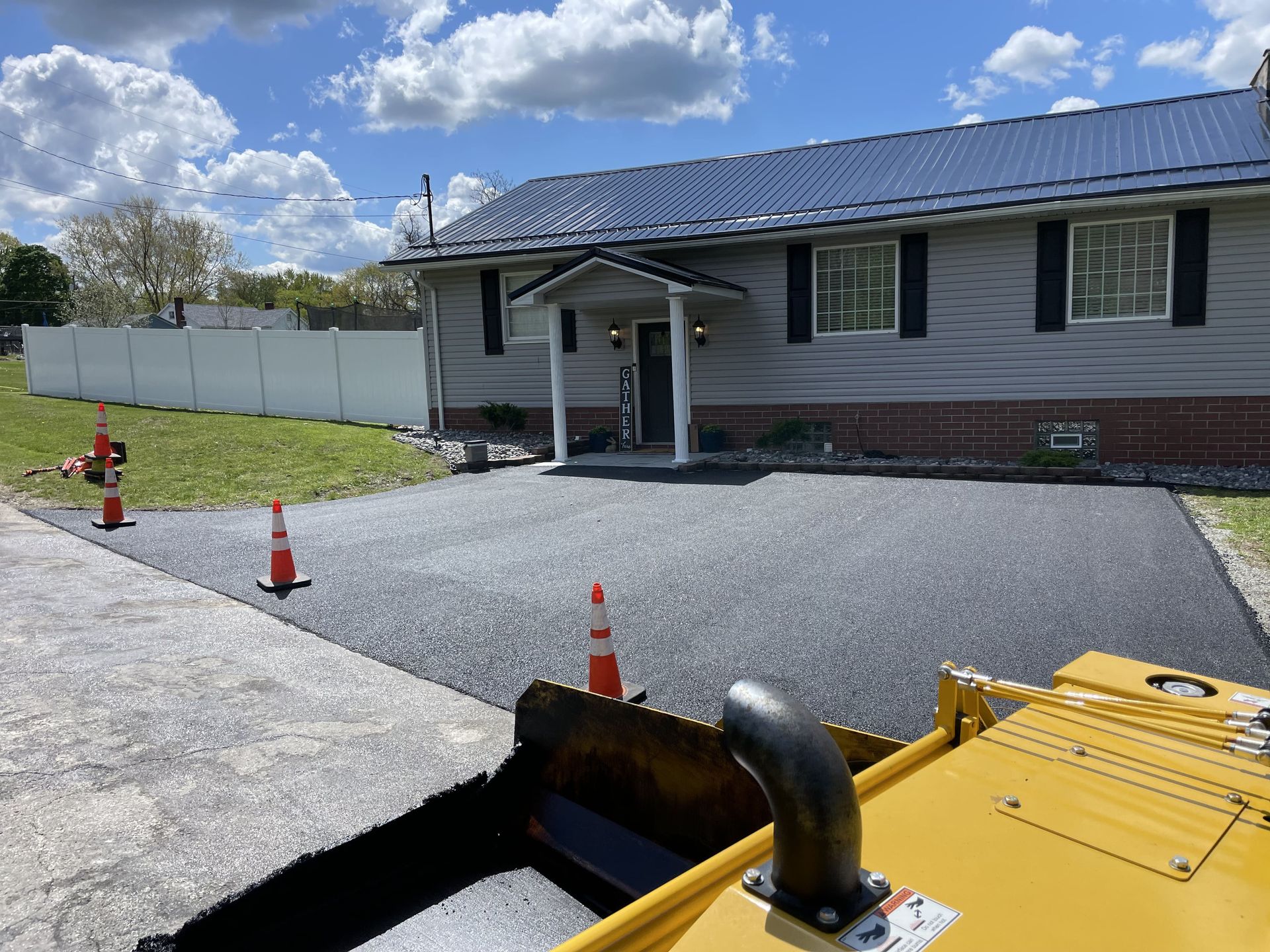 Asphalt paving in front of a gray house with black shutters, orange traffic cones, blue sky.