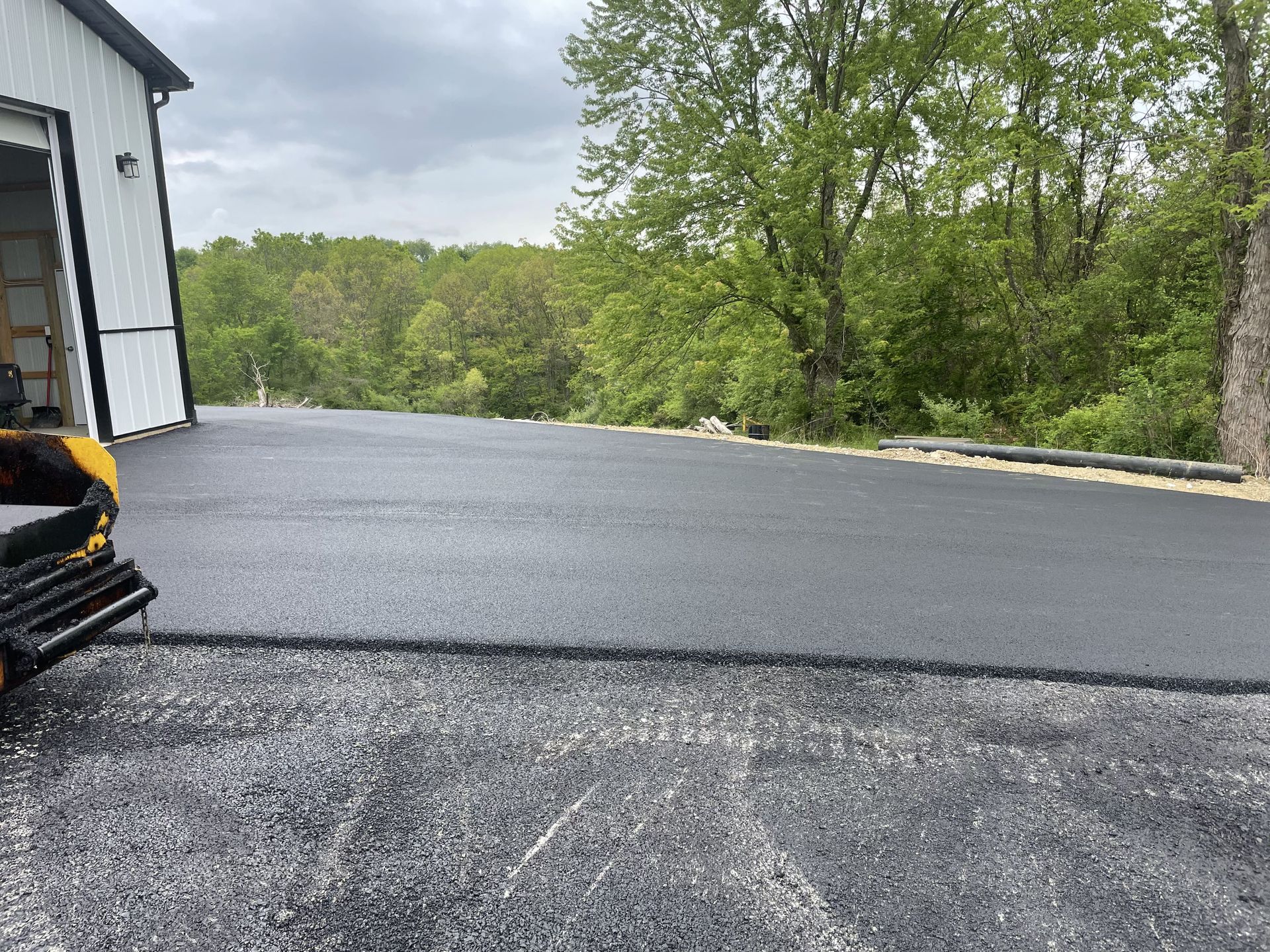 Newly paved asphalt driveway next to a white building with trees in the background.