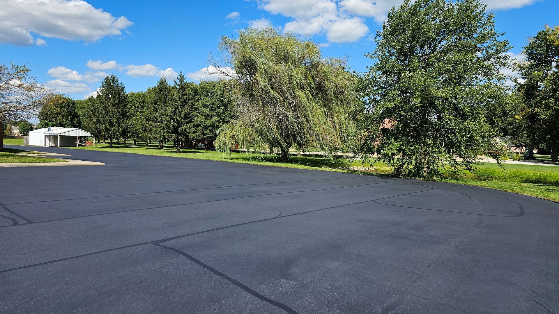 Paved parking lot beside green trees under a bright blue sky with scattered clouds