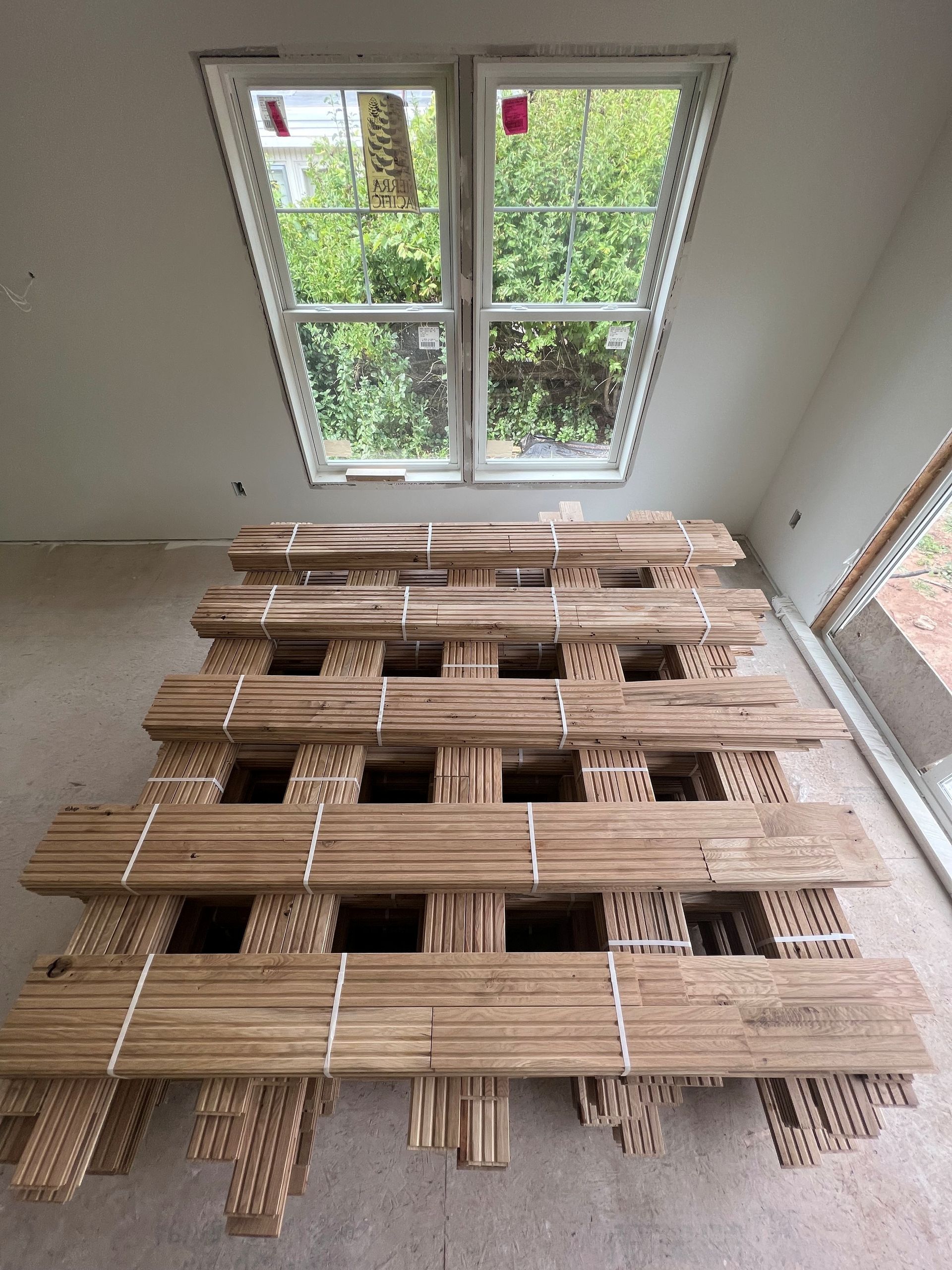 A bundle of wood flooring planks stacked in front of a window, inside a room under construction.