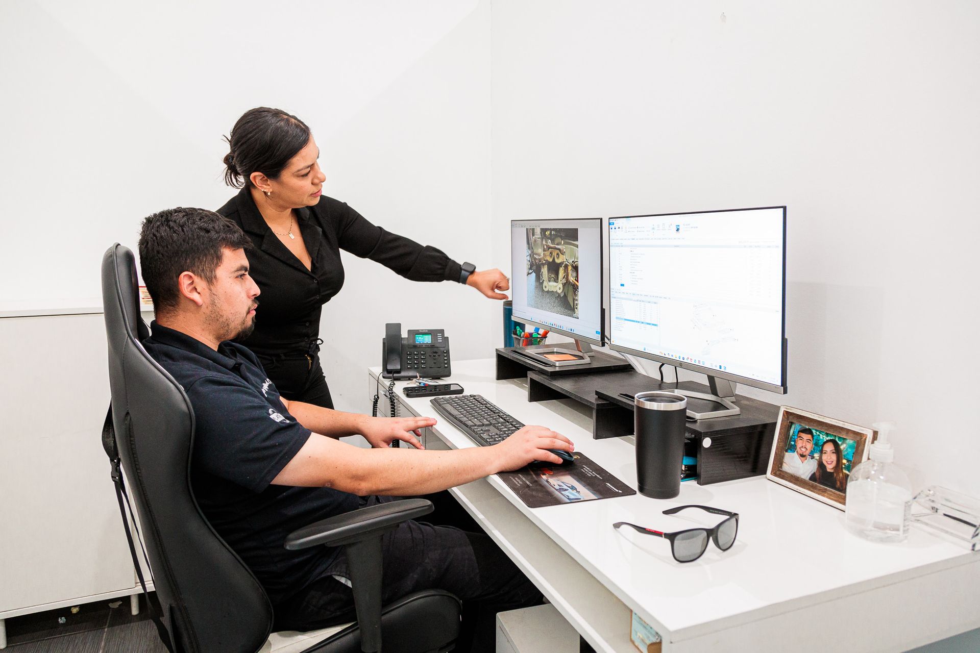 A woman is standing next to a man sitting at a desk using a computer.