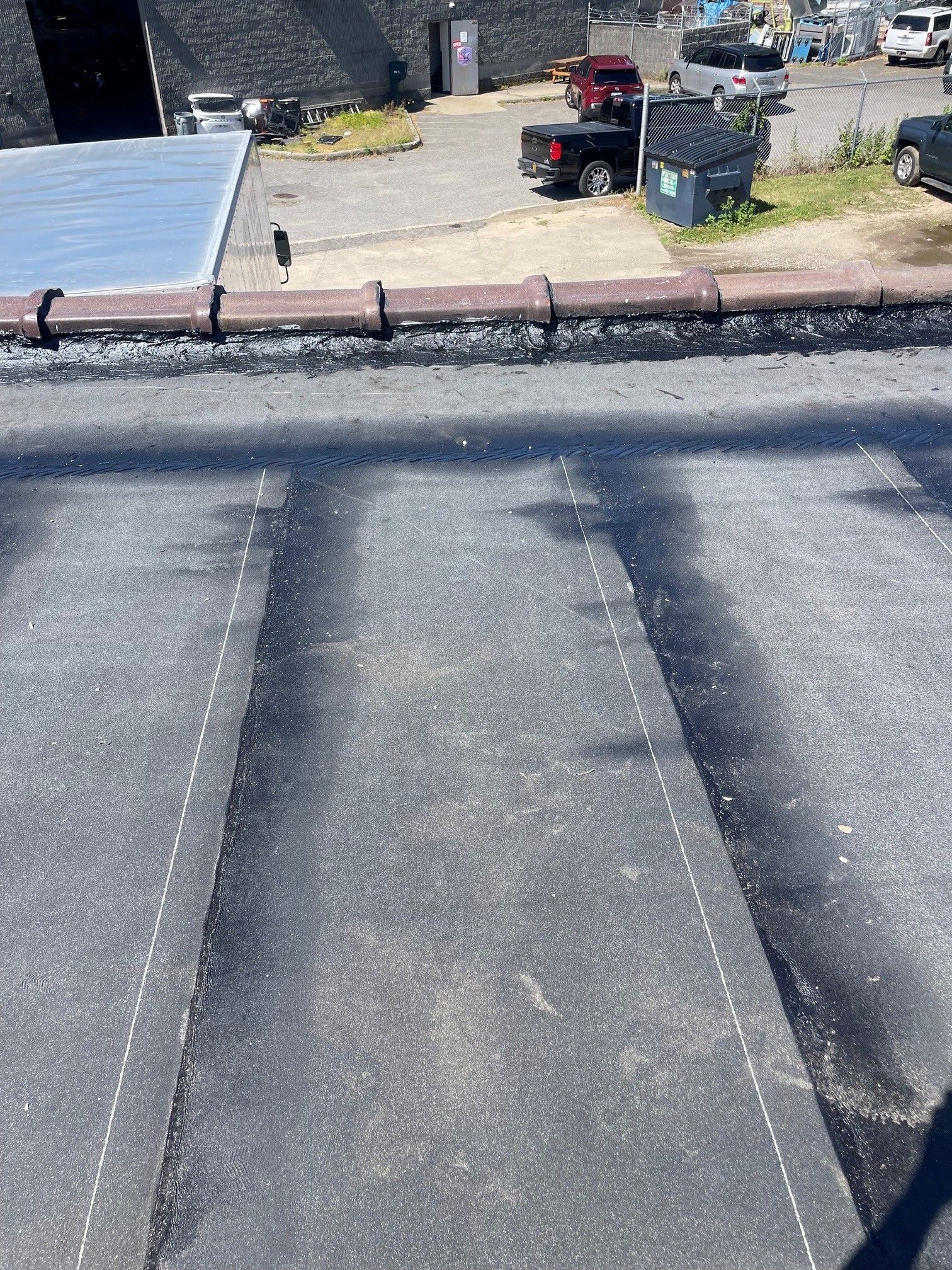 Rooftop view of a flat gravel roof with shadows, a brick edge, and a yard with tools and equipment beyond it