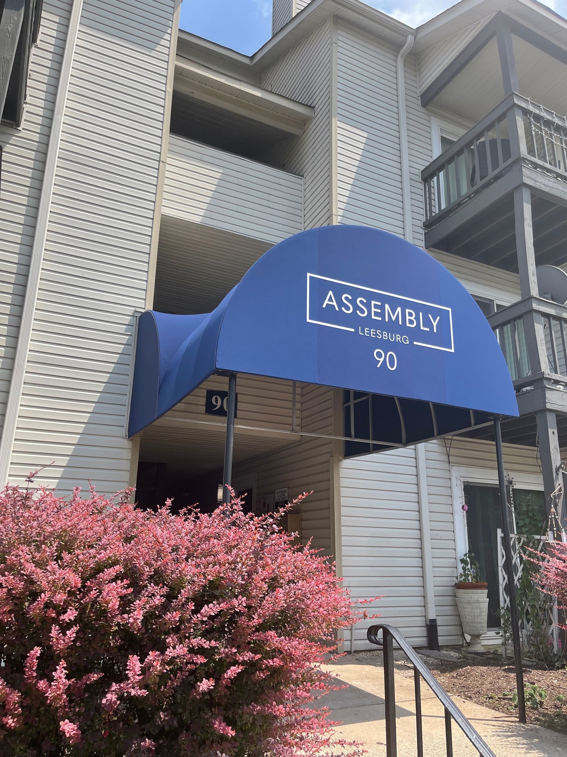 Blue awning at apartment entrance labeled “Assembly,” with pink flowering bushes and stairs.