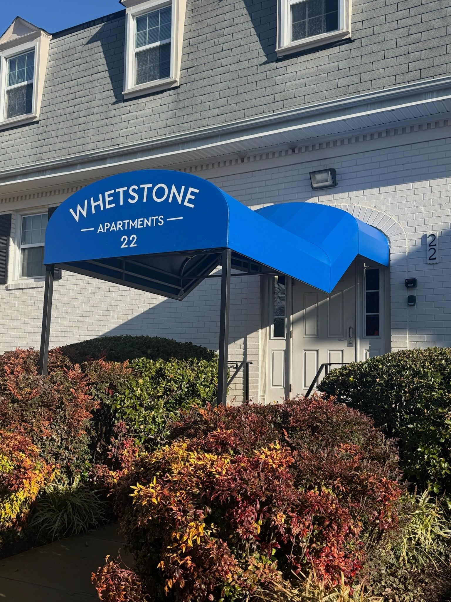 Blue awning over building entrance reading “WHETSTONE 32,” with shrubs in front.