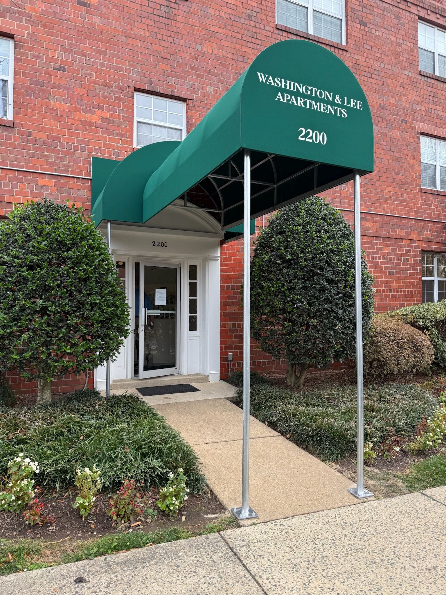 Green awning over apartment entrance with brick facade, walkway, and trimmed shrubs.