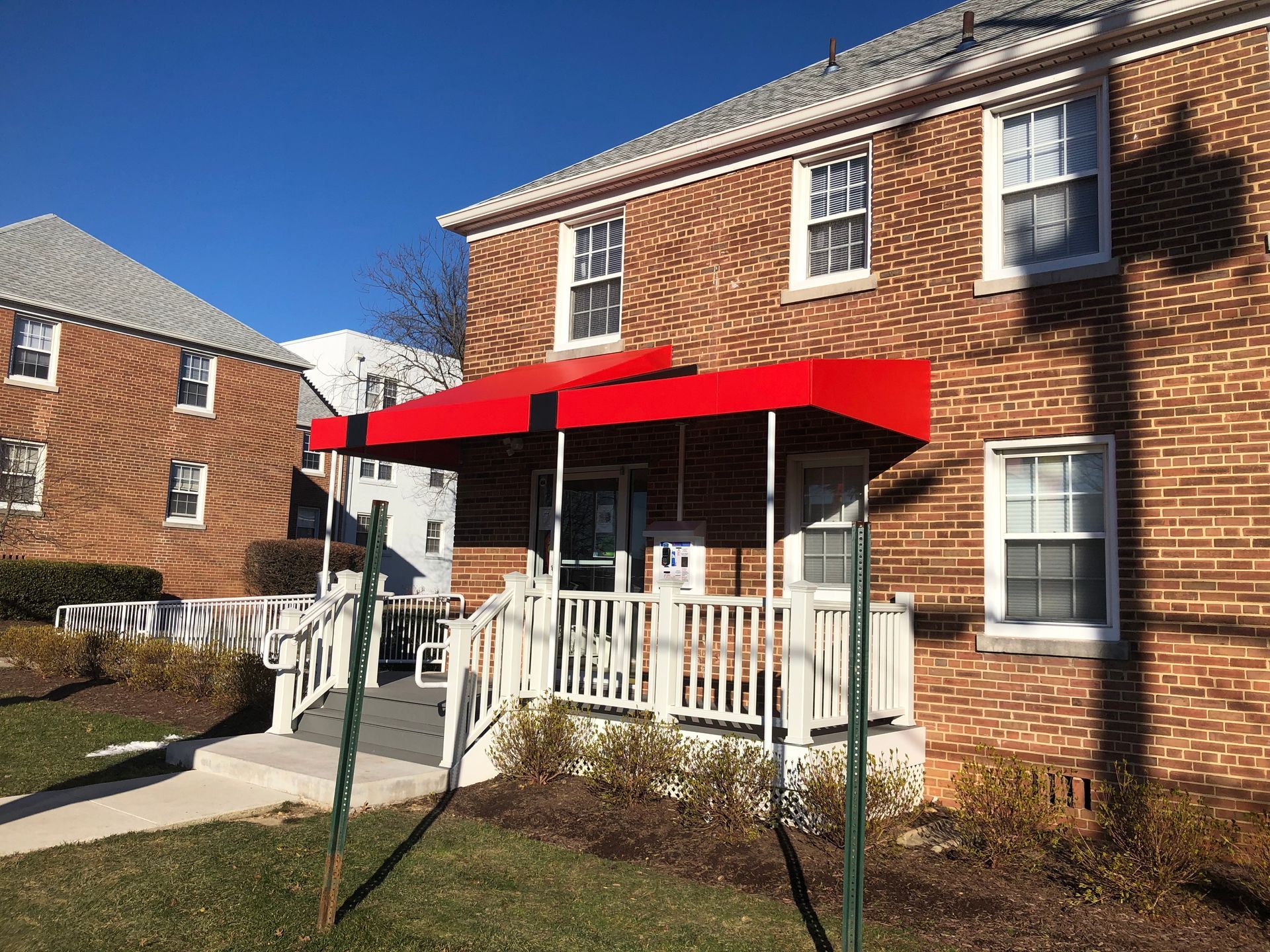 Brick apartment building with a red awning over a white porch railing on a sunny day.