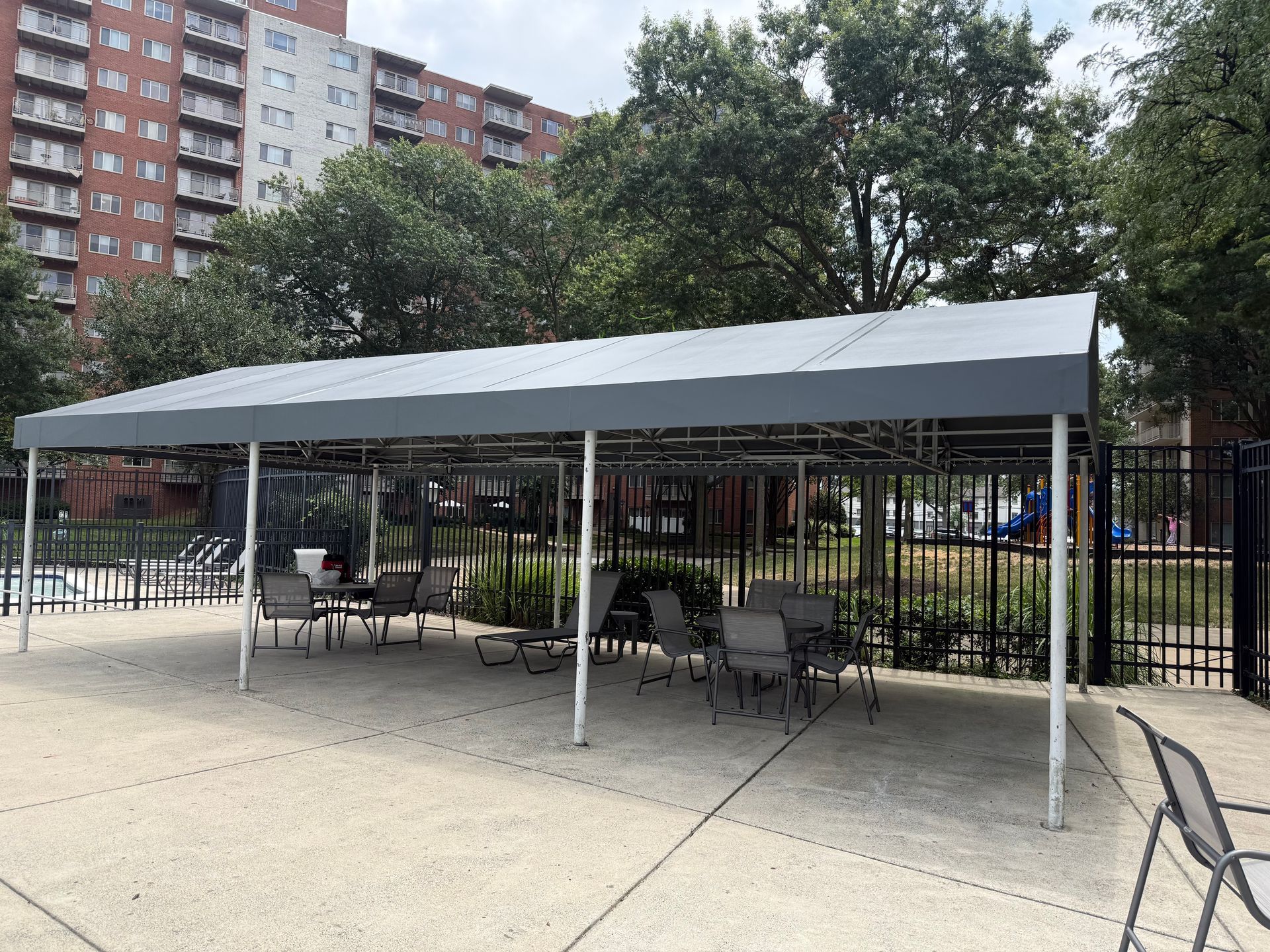 Covered outdoor seating area with tables and chairs beside a fenced courtyard and apartment buildings.