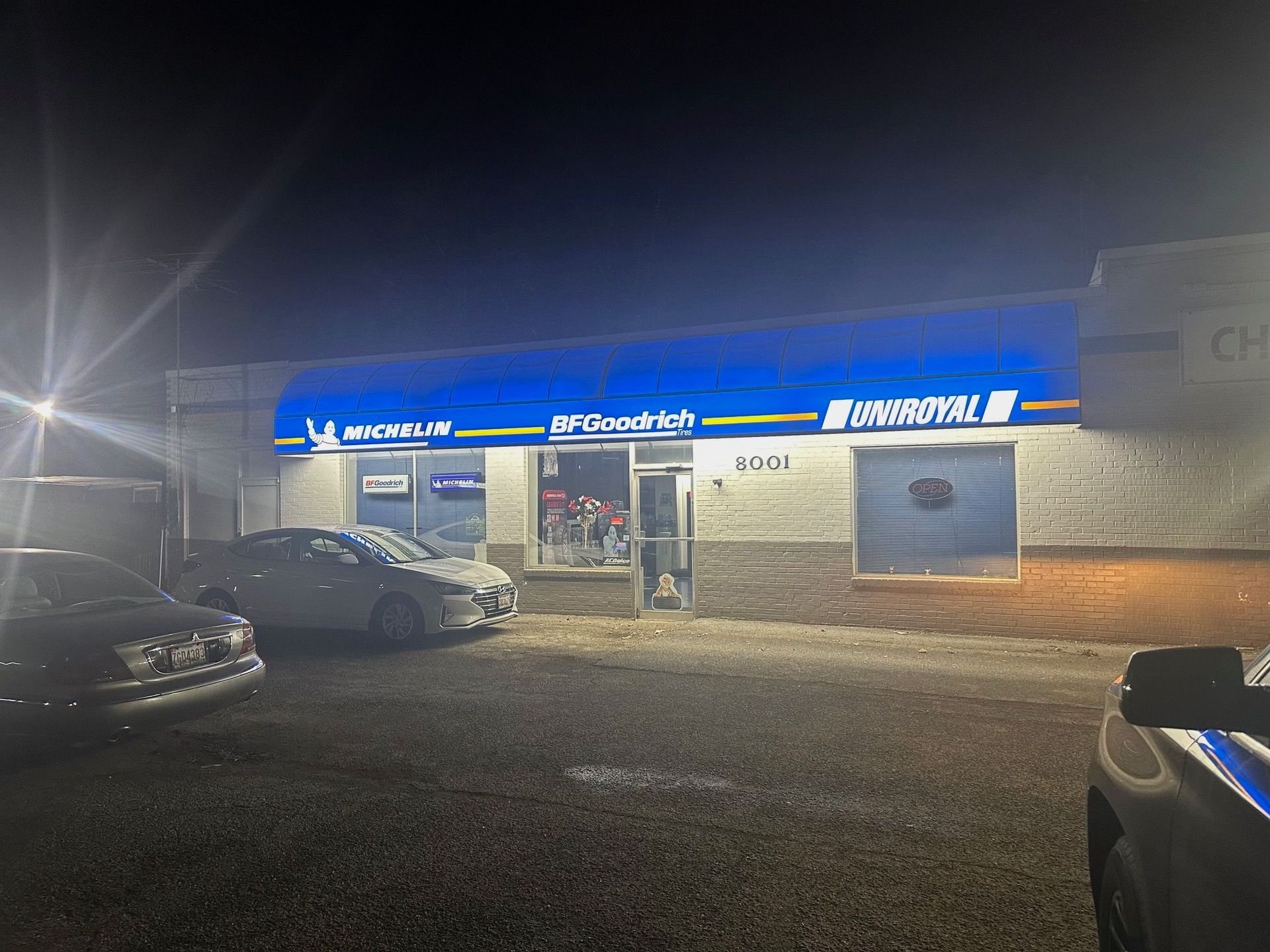 Nighttime view of a blue-lit storefront with parked cars in front and bright lights spilling onto the sidewalk
