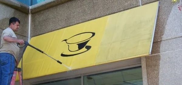 Man painting a yellow sign with a black hat and mustache logo above a storefront
