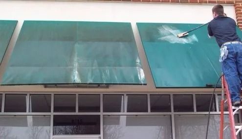 Worker cleaning a large green awning outside a building with a hose on a ladder