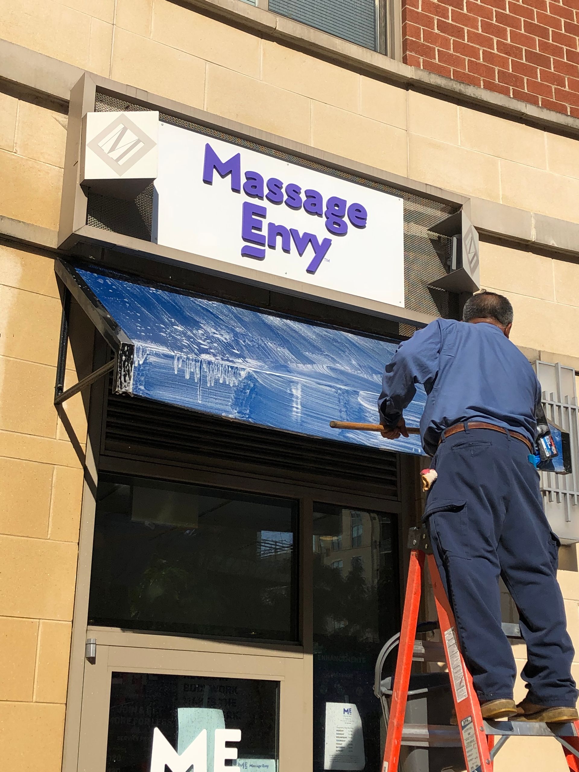 Worker on a ladder installing a blue awning at a storefront with the sign “Massage Envy”