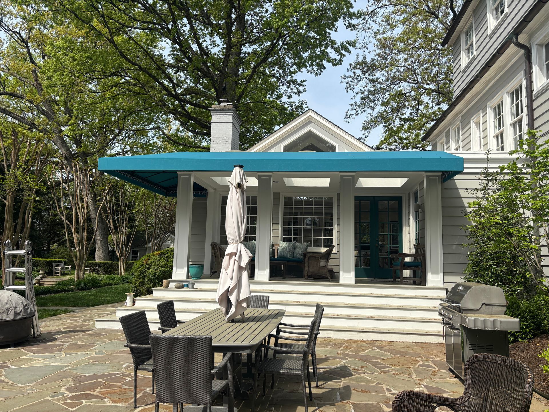 Patio with wicker dining table and chairs in front of a white house with blue awning and closed umbrellas