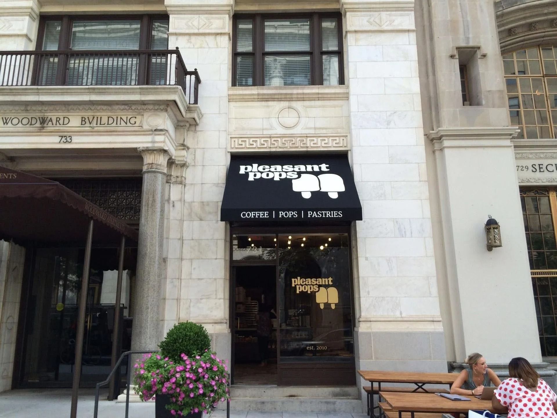 Storefront entrance with a black awning, potted flowers, and two people seated at an outdoor table nearby.