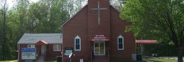 Small red brick church with white crosses, a sign by the entrance, and trees in the background