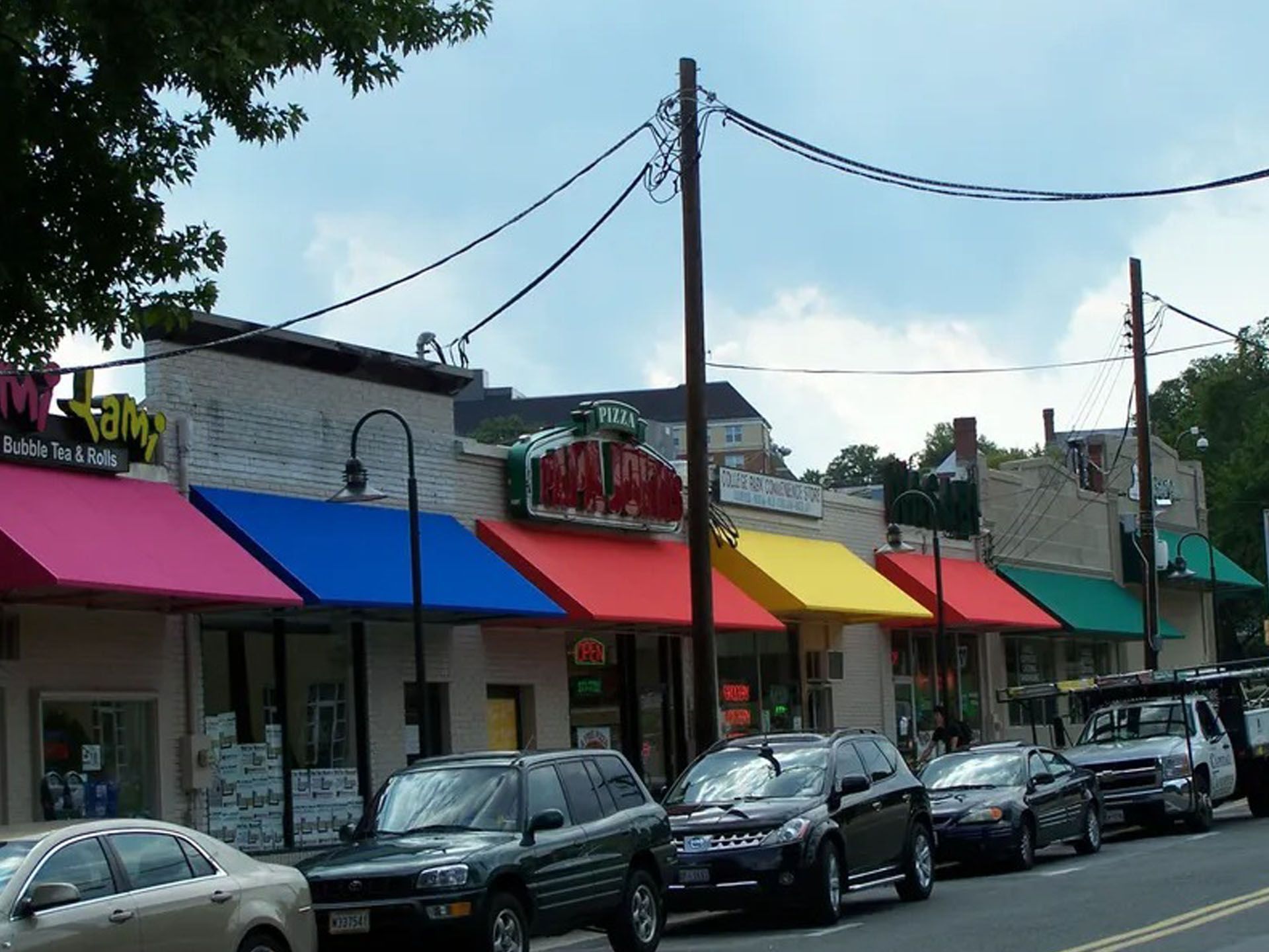 Row of storefronts with colorful awnings and parked cars along a street under utility poles