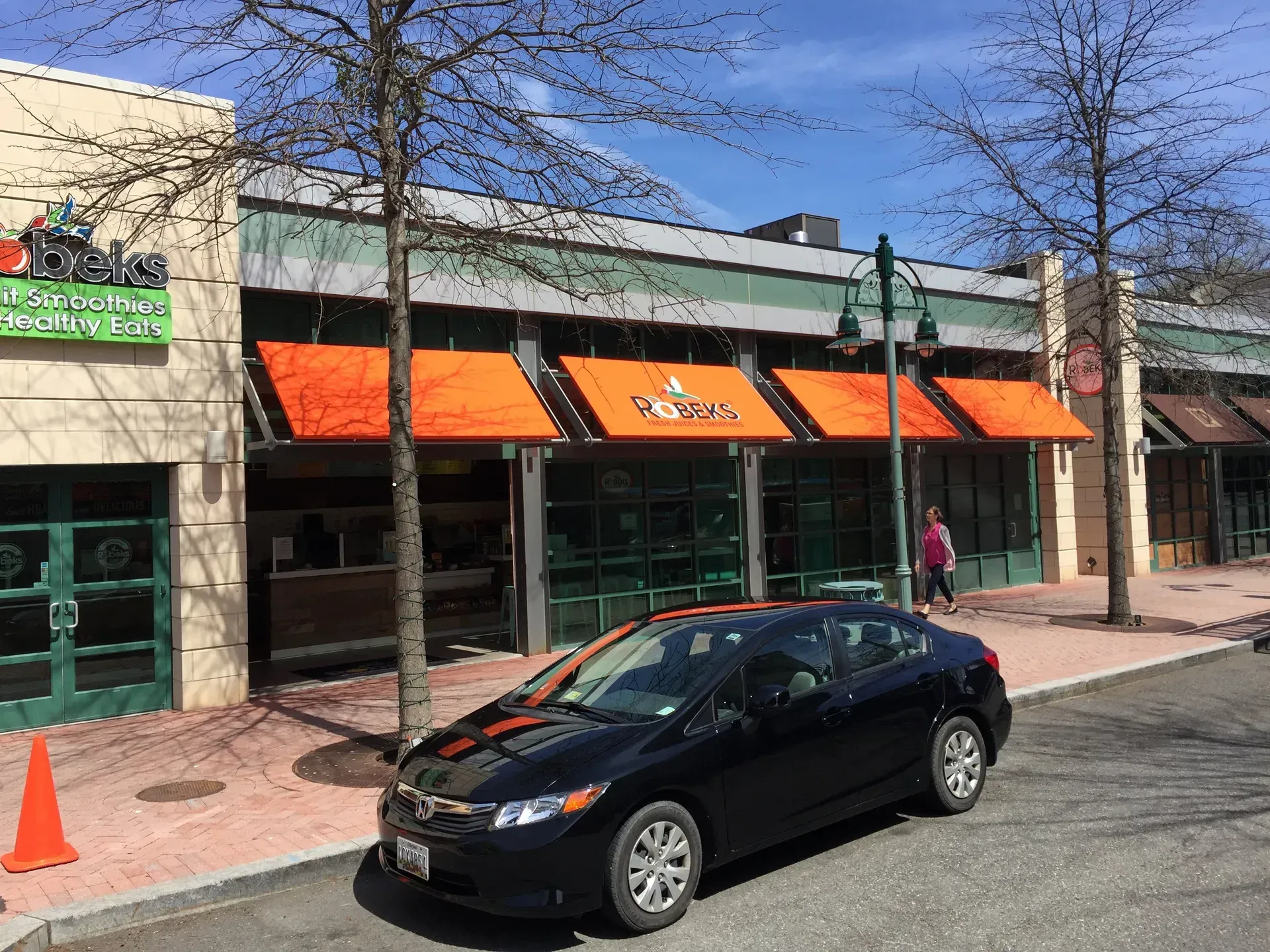 Street with storefronts and orange awnings, black car parked in front, and a pedestrian on the sidewalk.