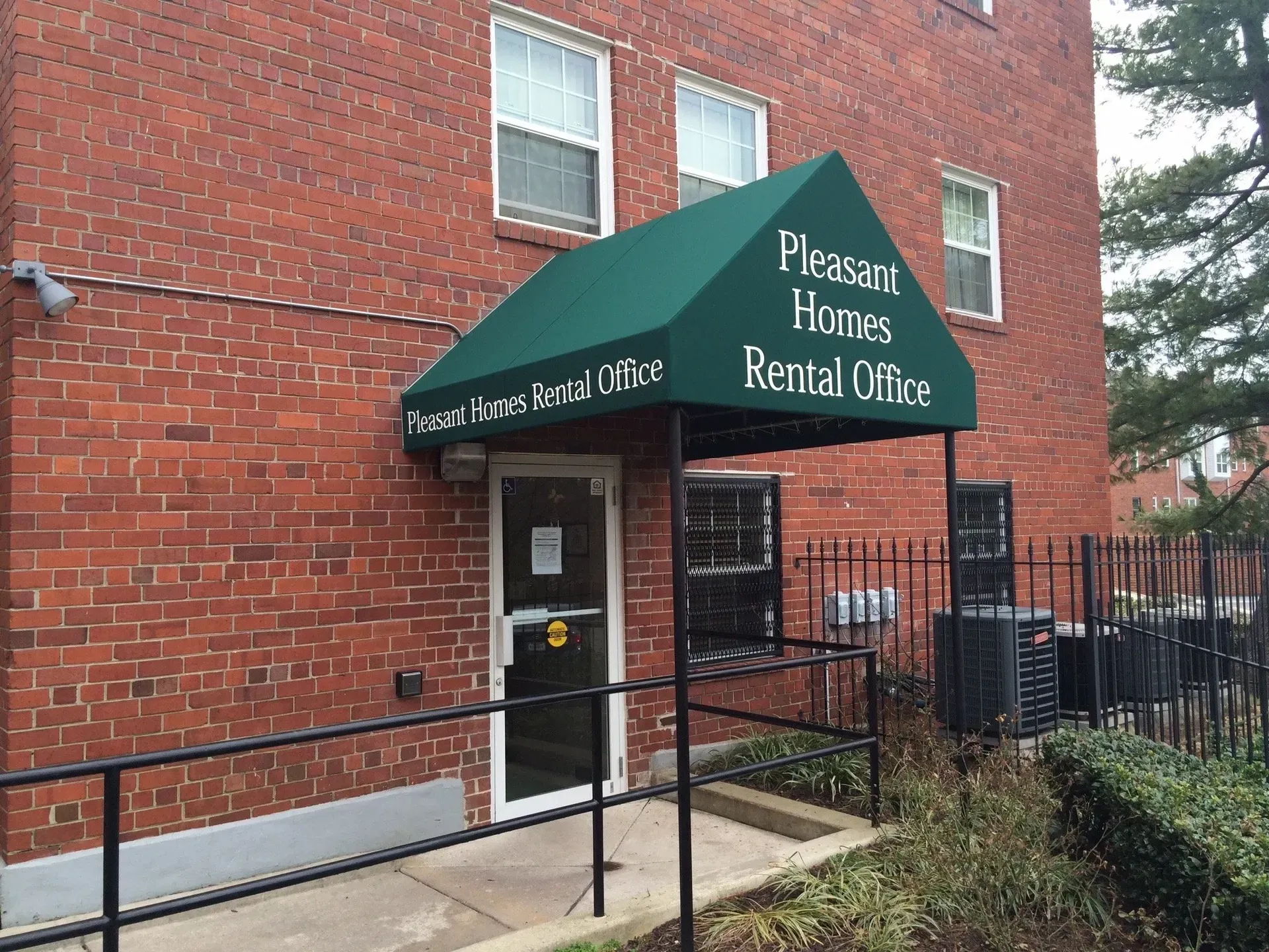 Pleasant Homes rental office entrance with green awning on a brick building, beside a ramp and black railing