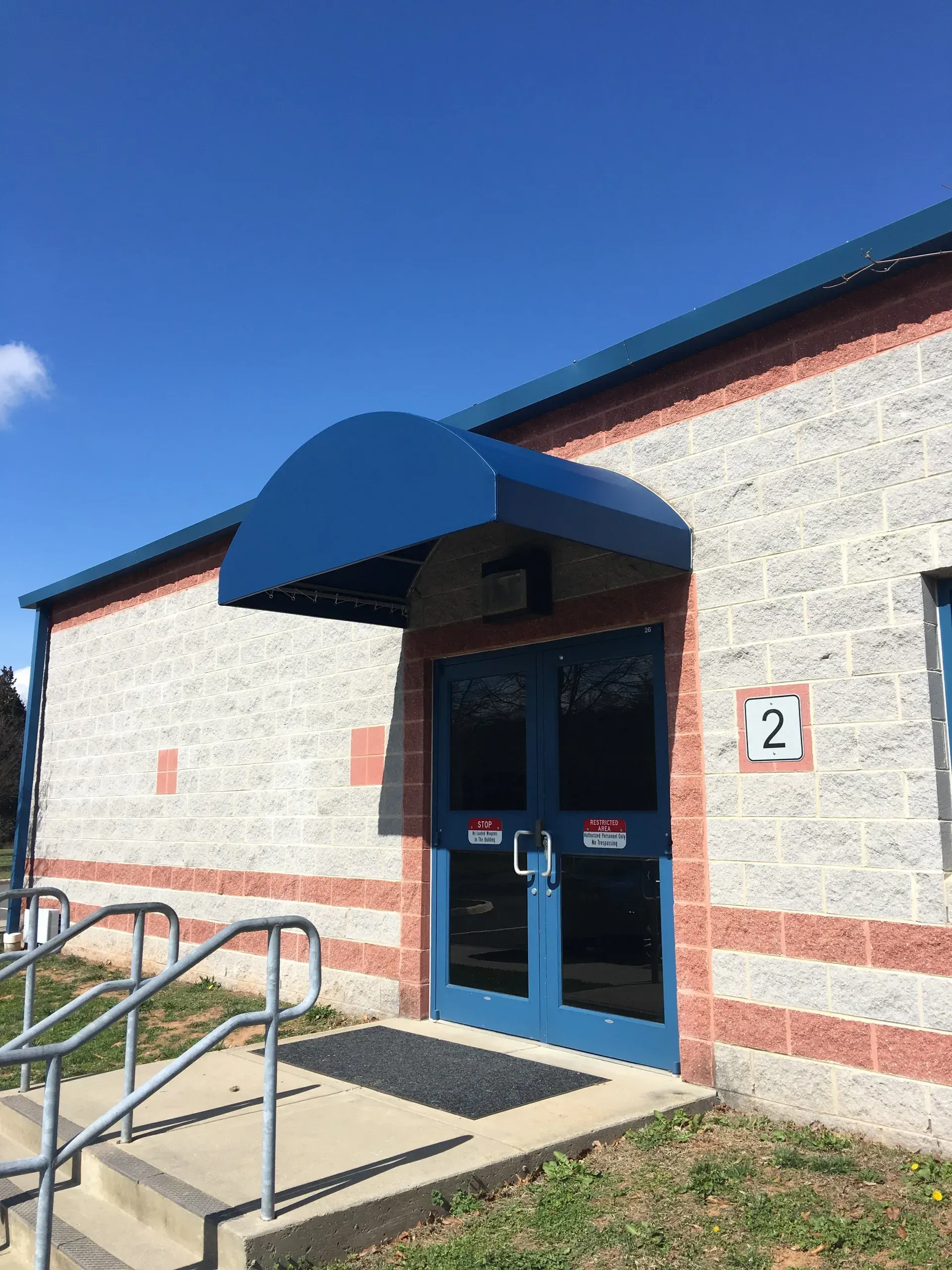 Brick building entrance with blue awning, double doors, and wheelchair ramp with handrails