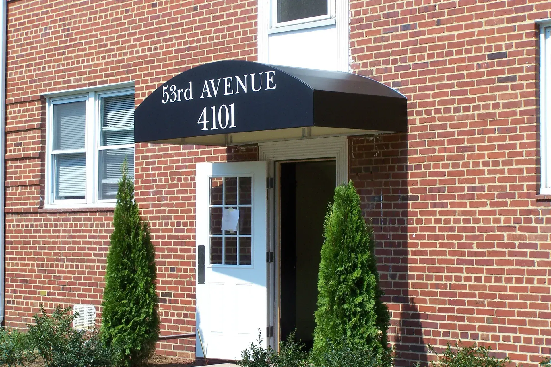 Brick building entrance with black awning reading “3rd Avenue 4101” and two shrubs flanking the door