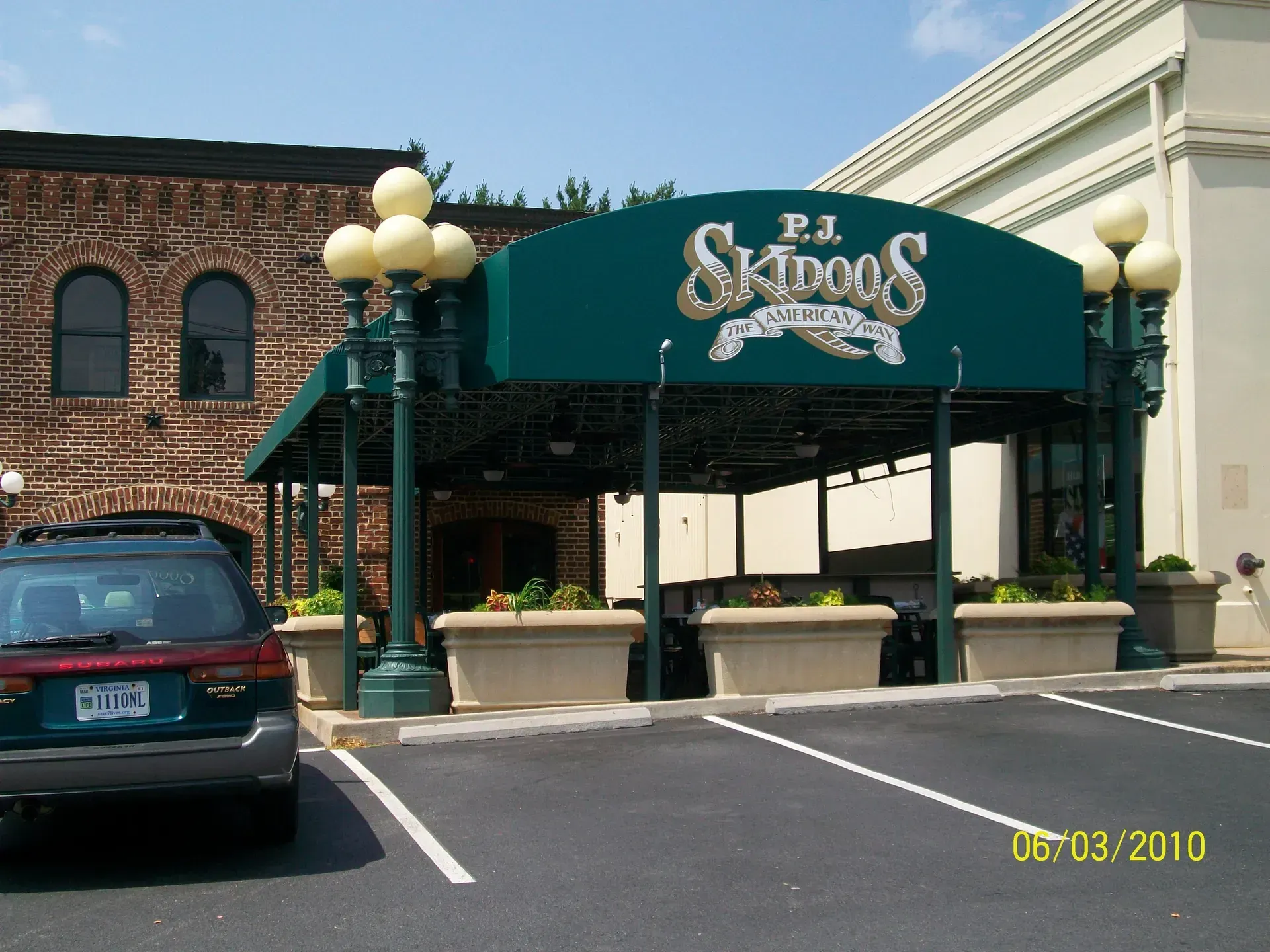 Skopos restaurant entrance with green awning, brick building, parked SUV, and outdoor planter boxes