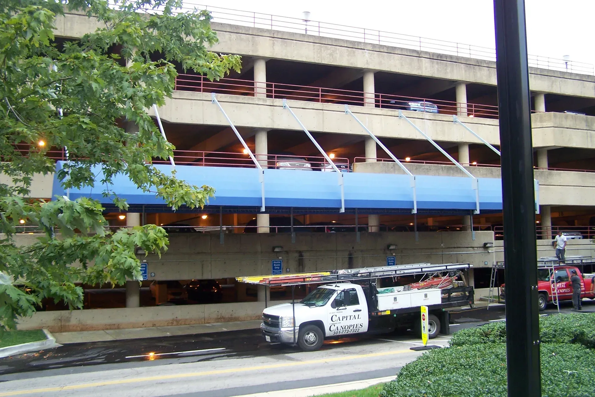 Multi-level parking garage with blue awning, parked vehicles, and trees along the street