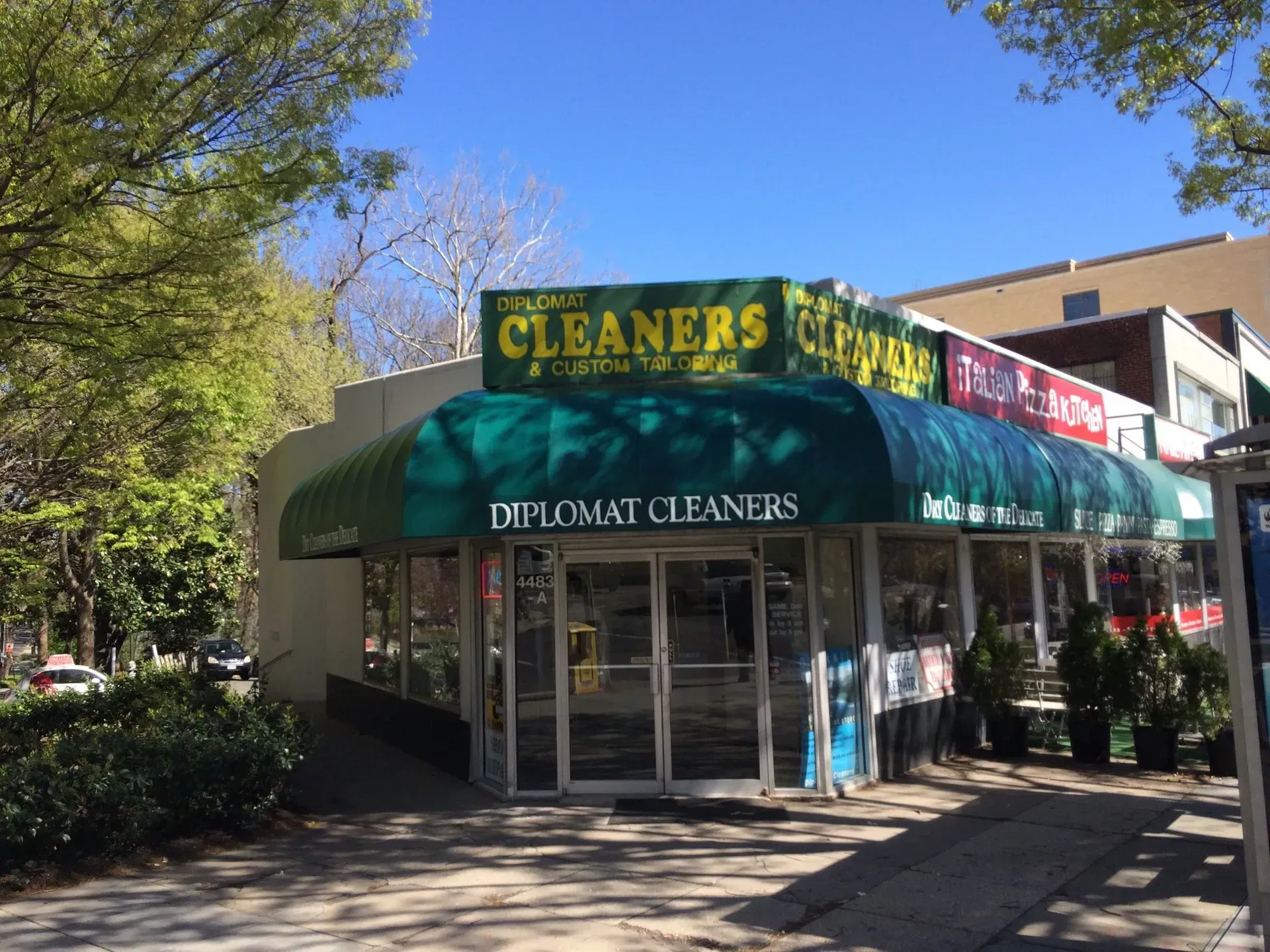 Corner storefront of Diplomat Cleaners with green awning on a sunny tree-lined street