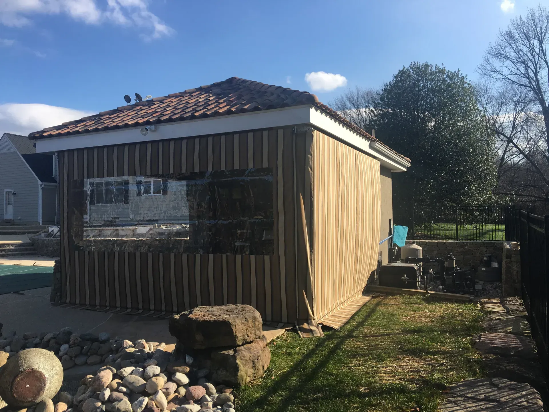 Small wooden shed with a tiled roof in a sunny yard, surrounded by rocks and a grassy path.