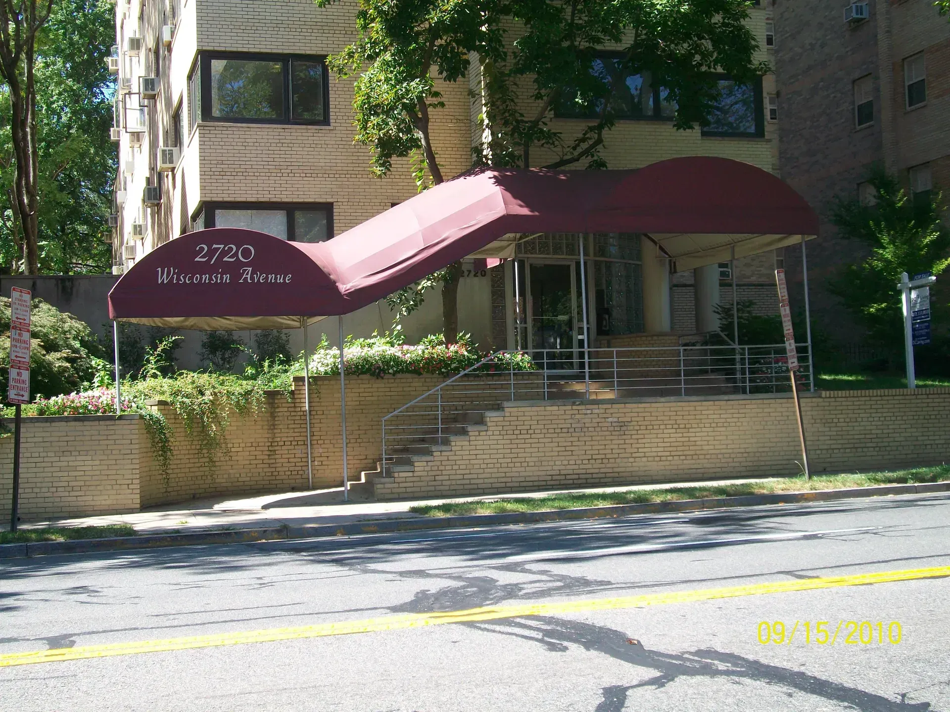 Apartment building entrance with burgundy awnings, steps, and greenery along a tree-lined street