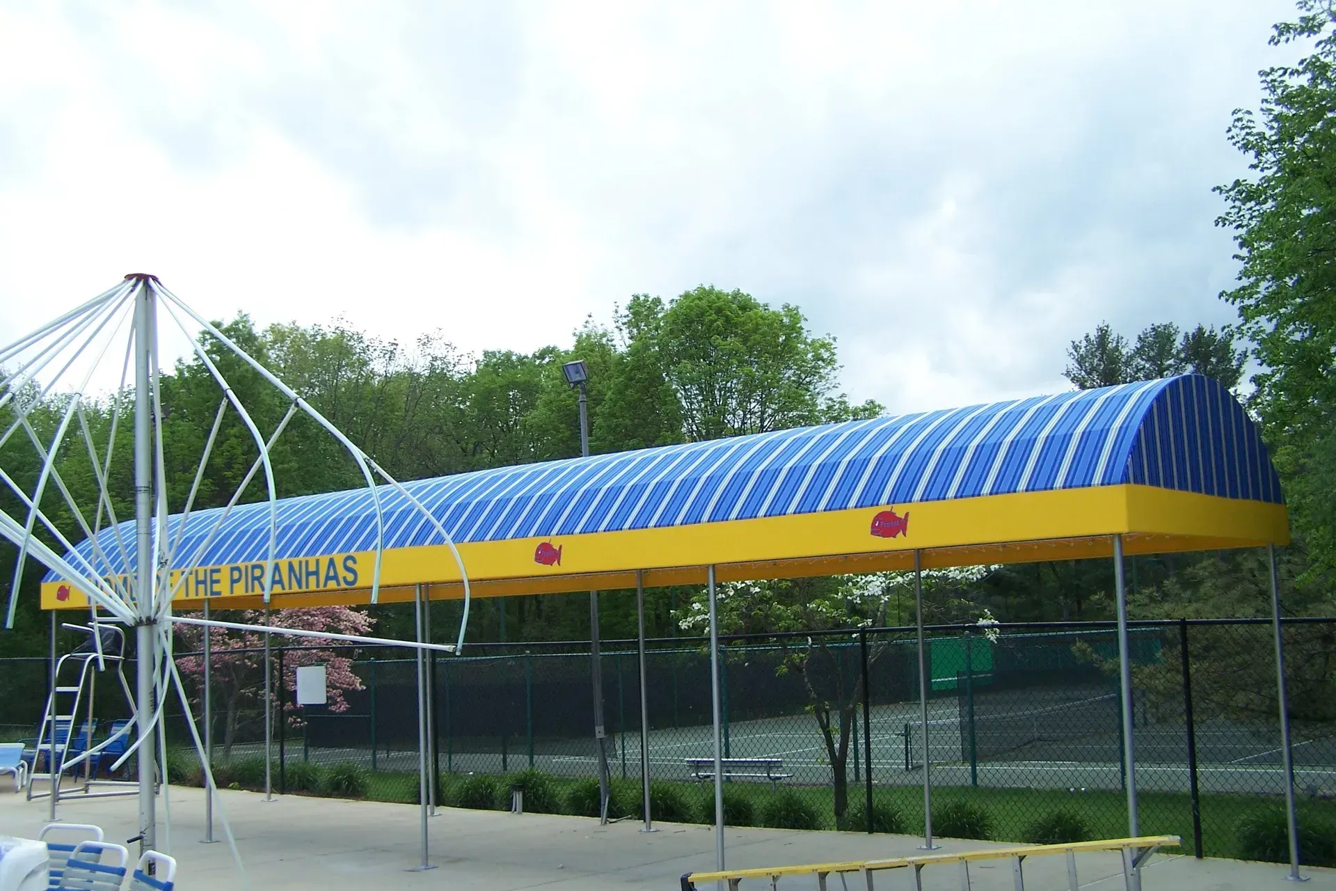 Blue-and-yellow canopy over a fenced outdoor sports court with trees in the background
