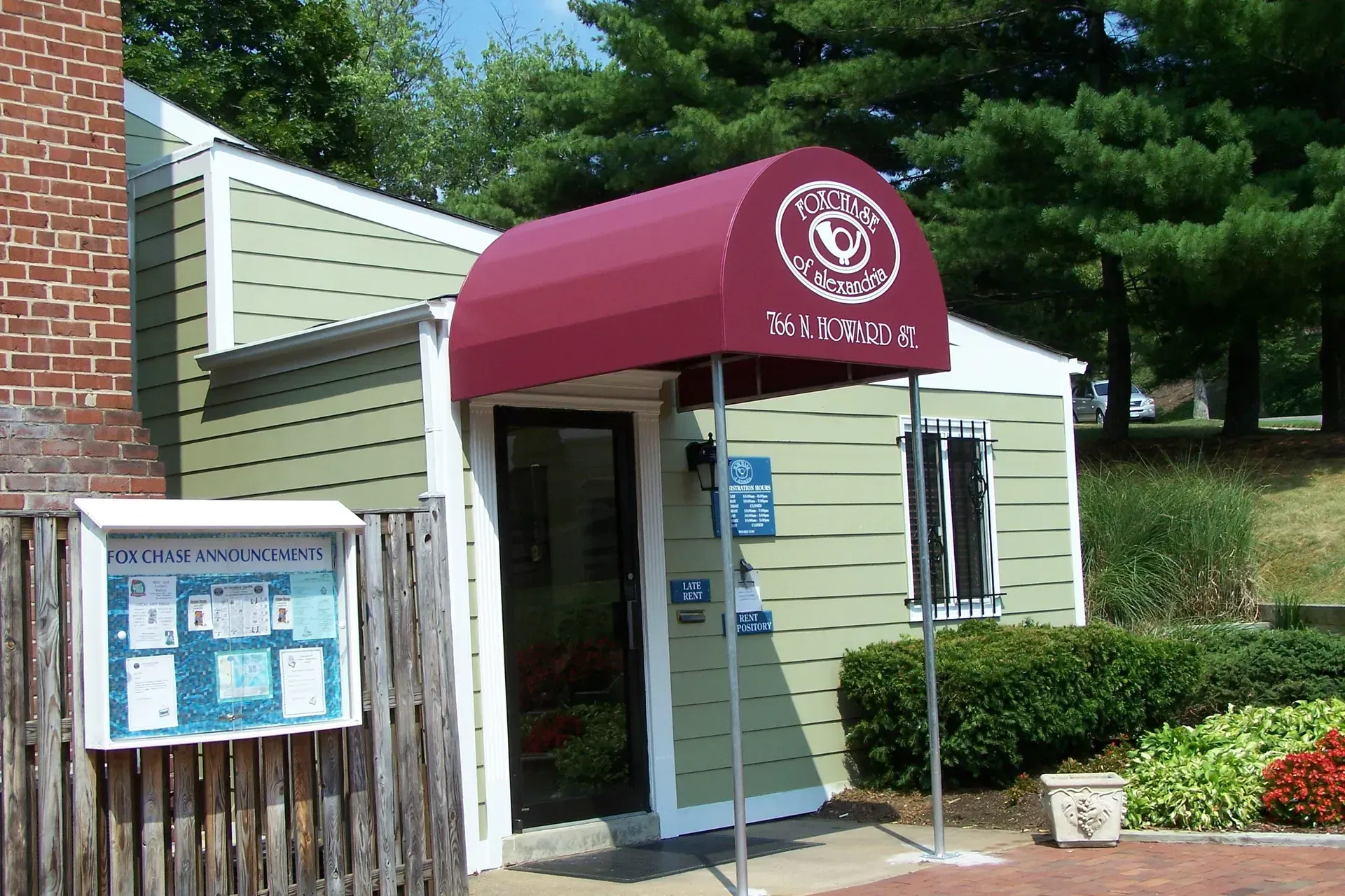 Entrance with maroon awning, glass door, and bulletin board outside a brick-and-siding building.