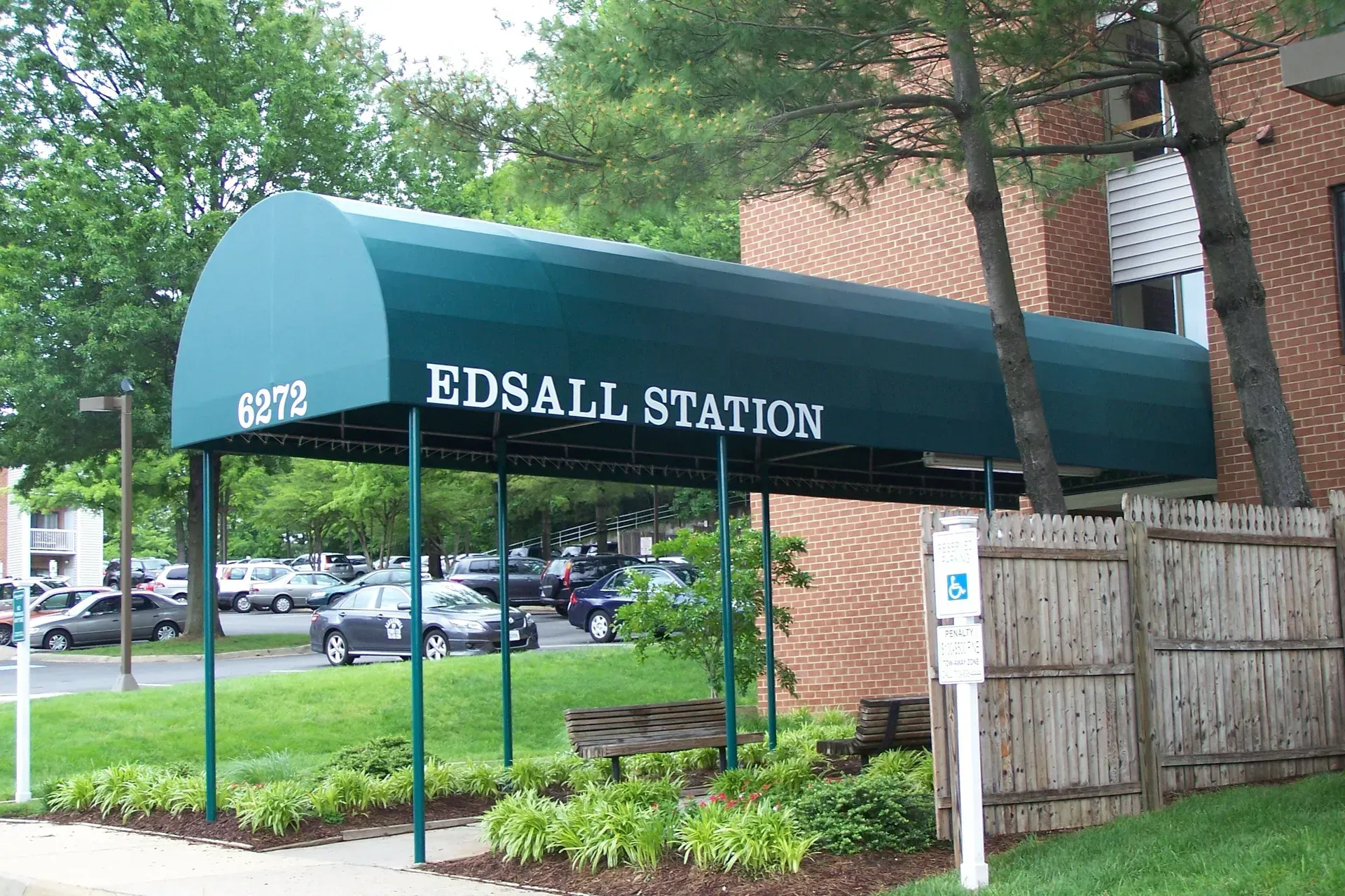 Edsall Station entrance with teal awning, wooden fence, and parked cars in front of a brick building.