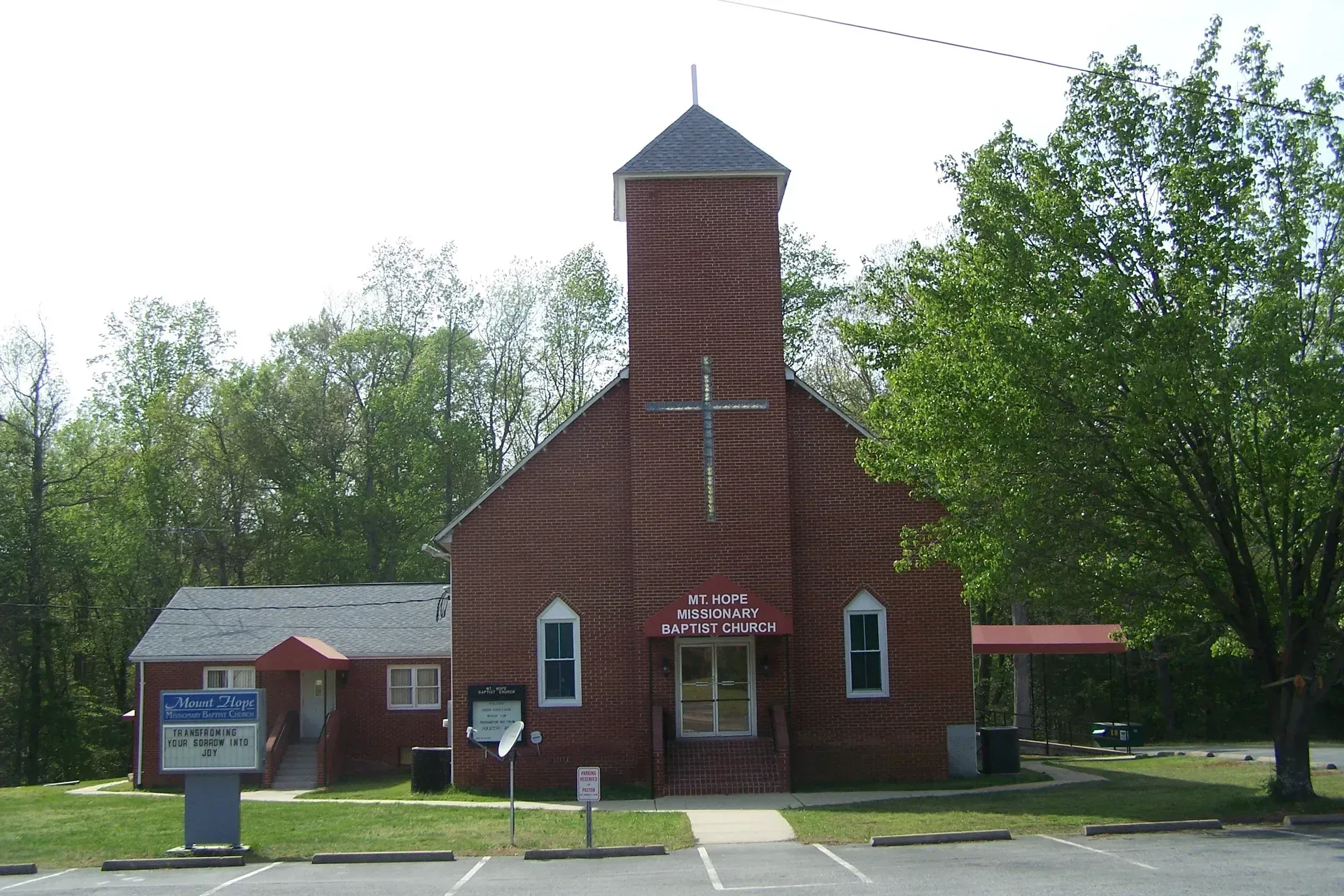 Small red brick church with a tower and sign, set beside a parking lot and trees.