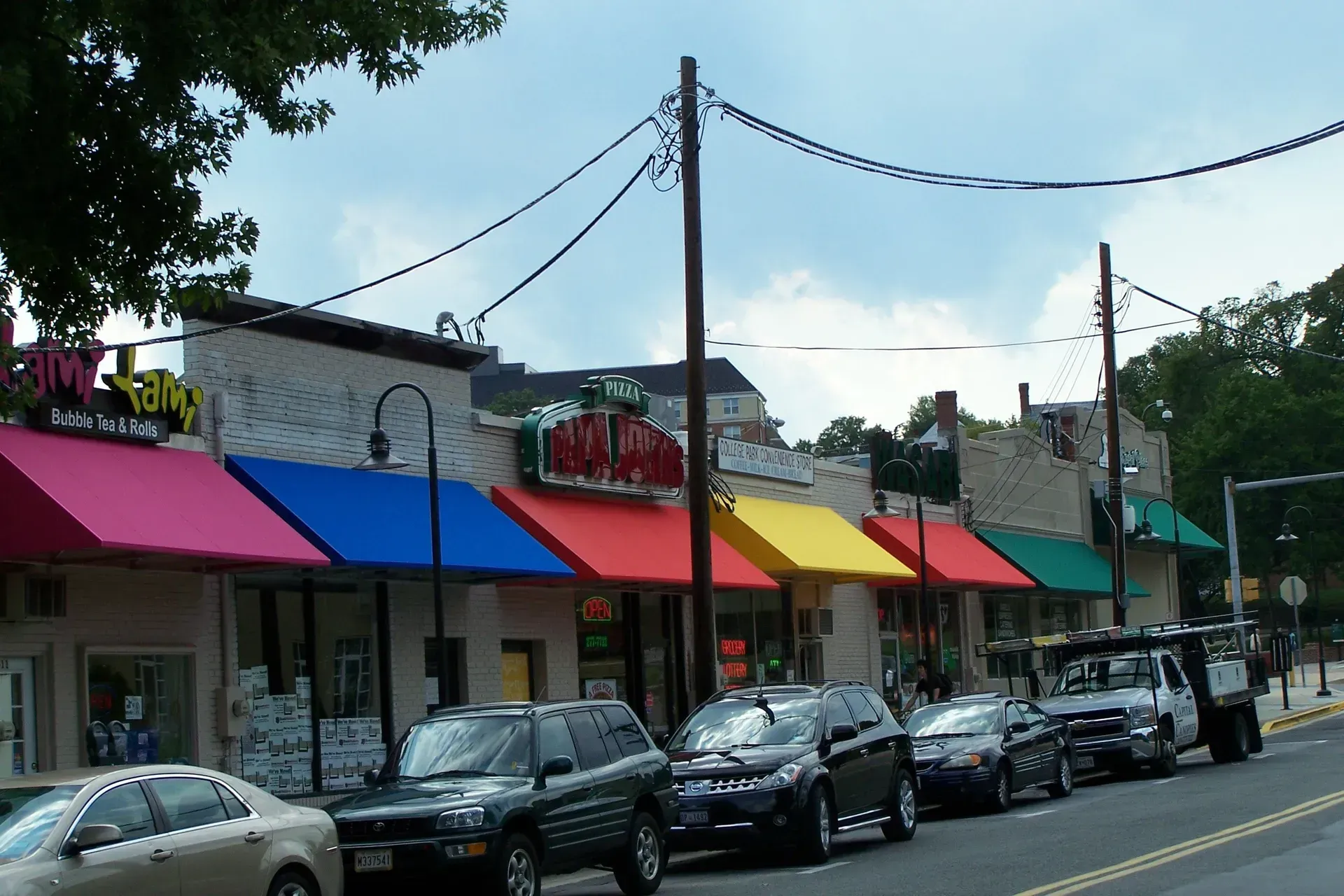 Colorful storefronts with pink, blue, red, yellow, and green awnings along a street with parked cars.