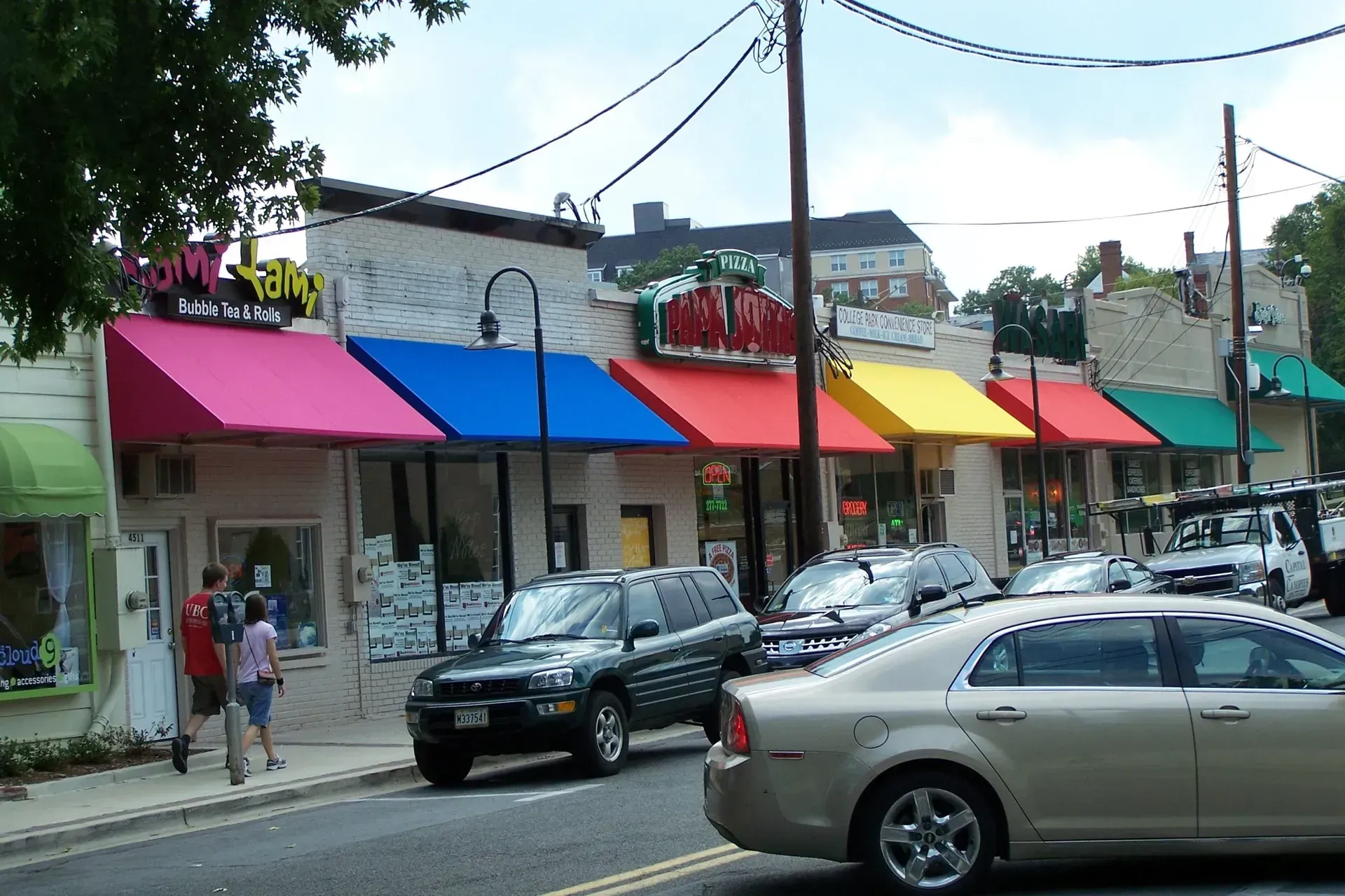 Colorful storefronts with awnings on a busy street, with cars parked and pedestrians walking by.