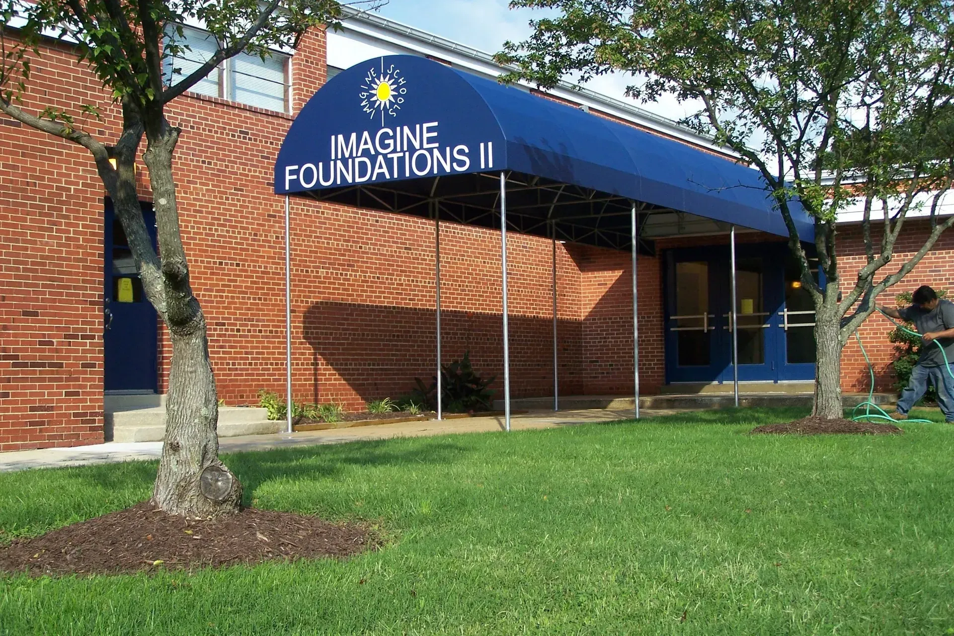 Brick building with blue awning reading “Imagine Foundations II” beside a grassy lawn and tree
