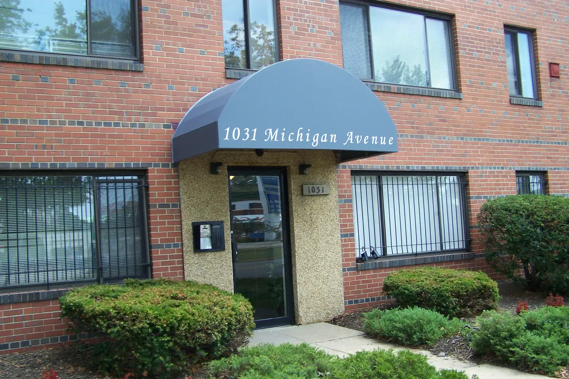 Brick apartment building entrance with glass door, blue awning, and shrubs outside