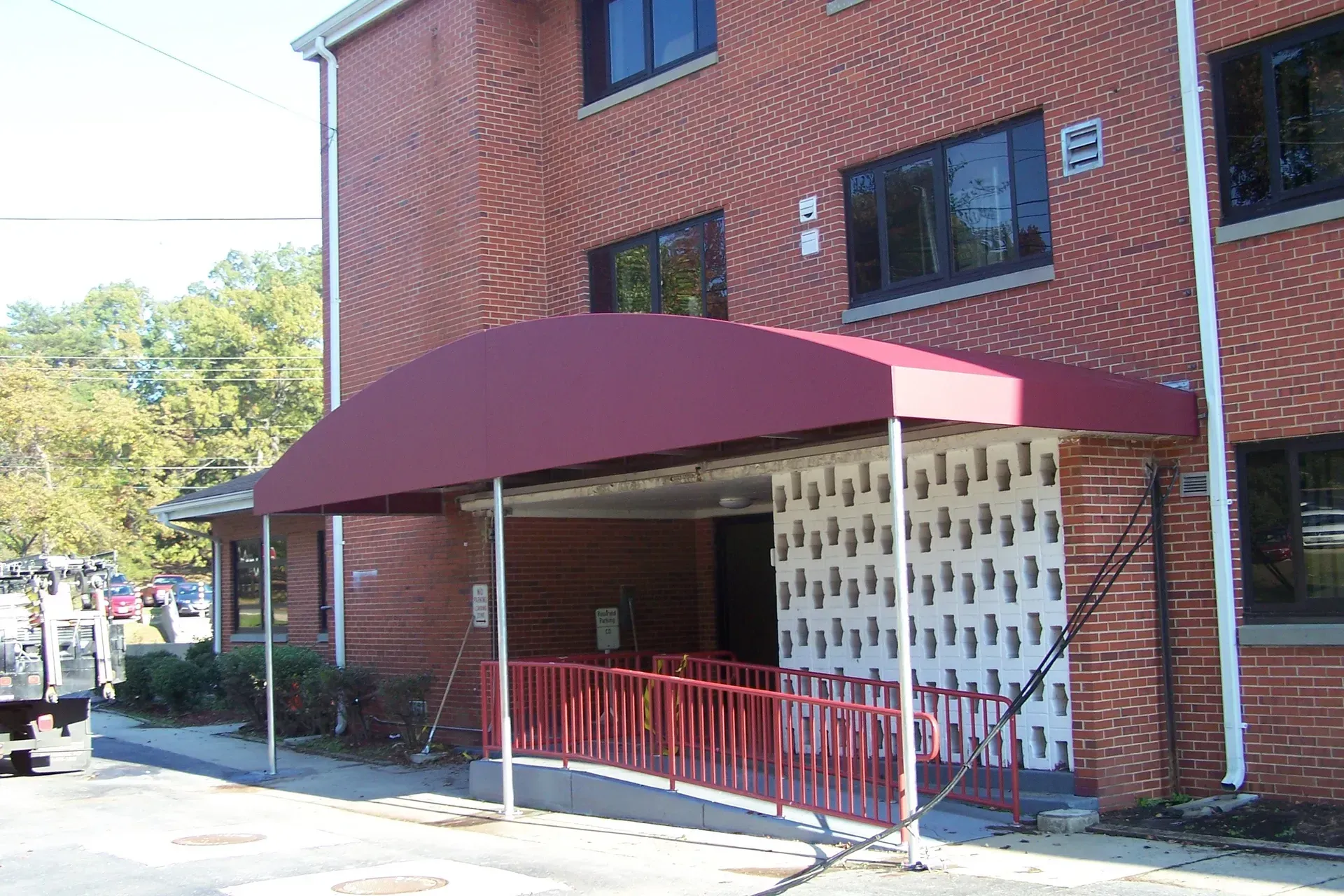 Brick building entrance with maroon awning and red railing on a sunny street.