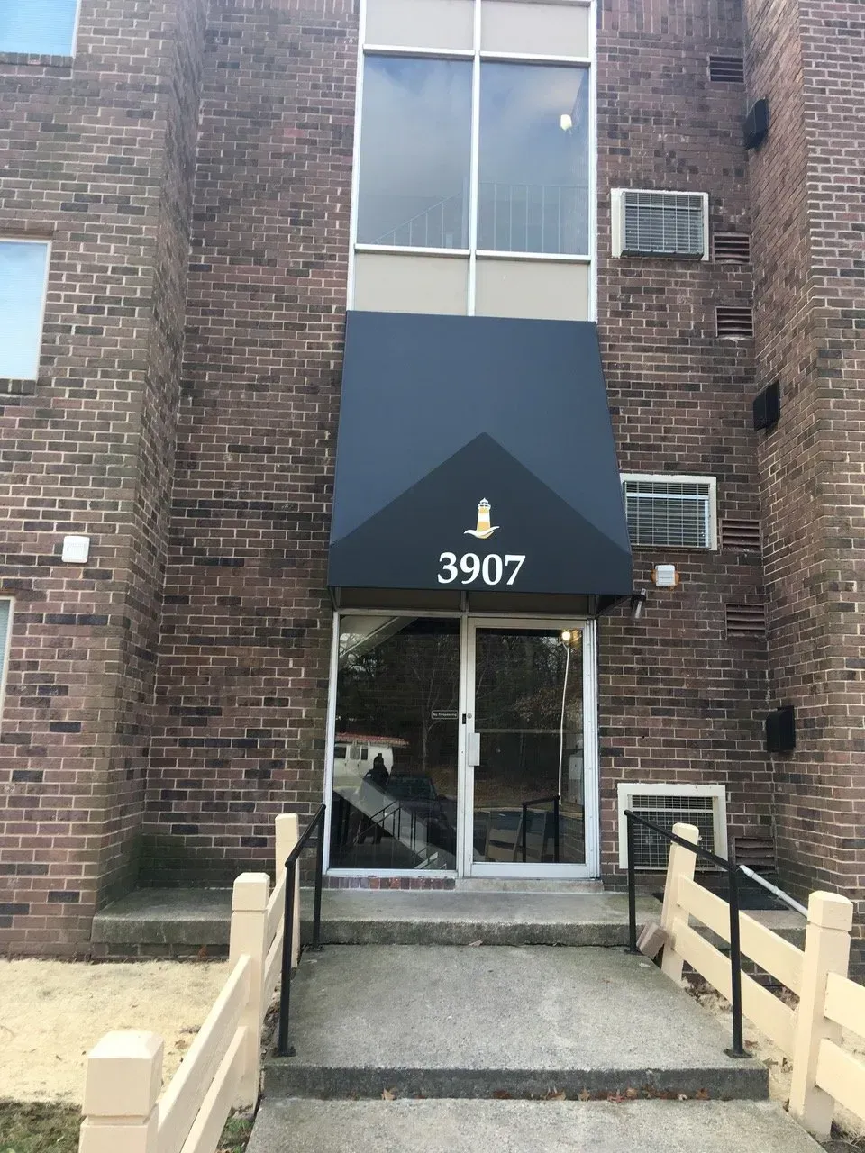 Brick apartment entrance with black awning marked 3907 and a ramp leading to glass doors