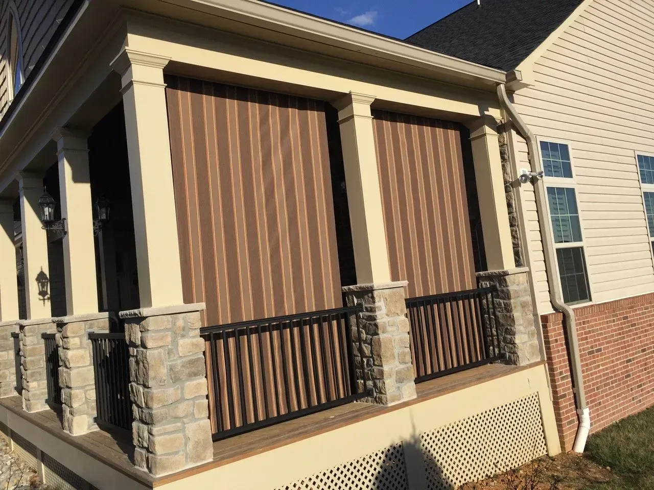 Covered porch with stone columns, brown blinds, and black railing on a beige house.