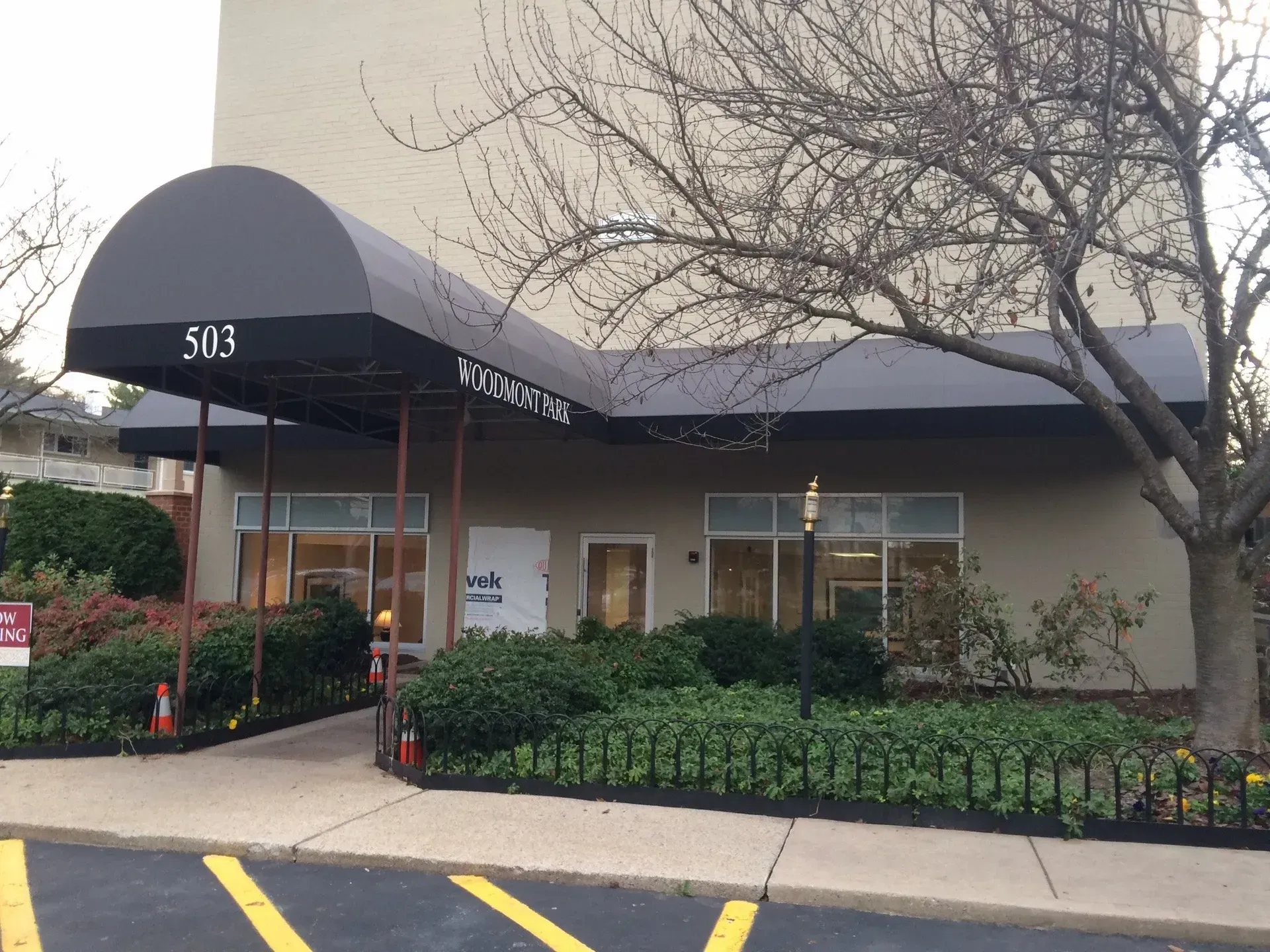 Exterior of a building with a black awning, shrubs, and a leafless tree by a parking lot entrance