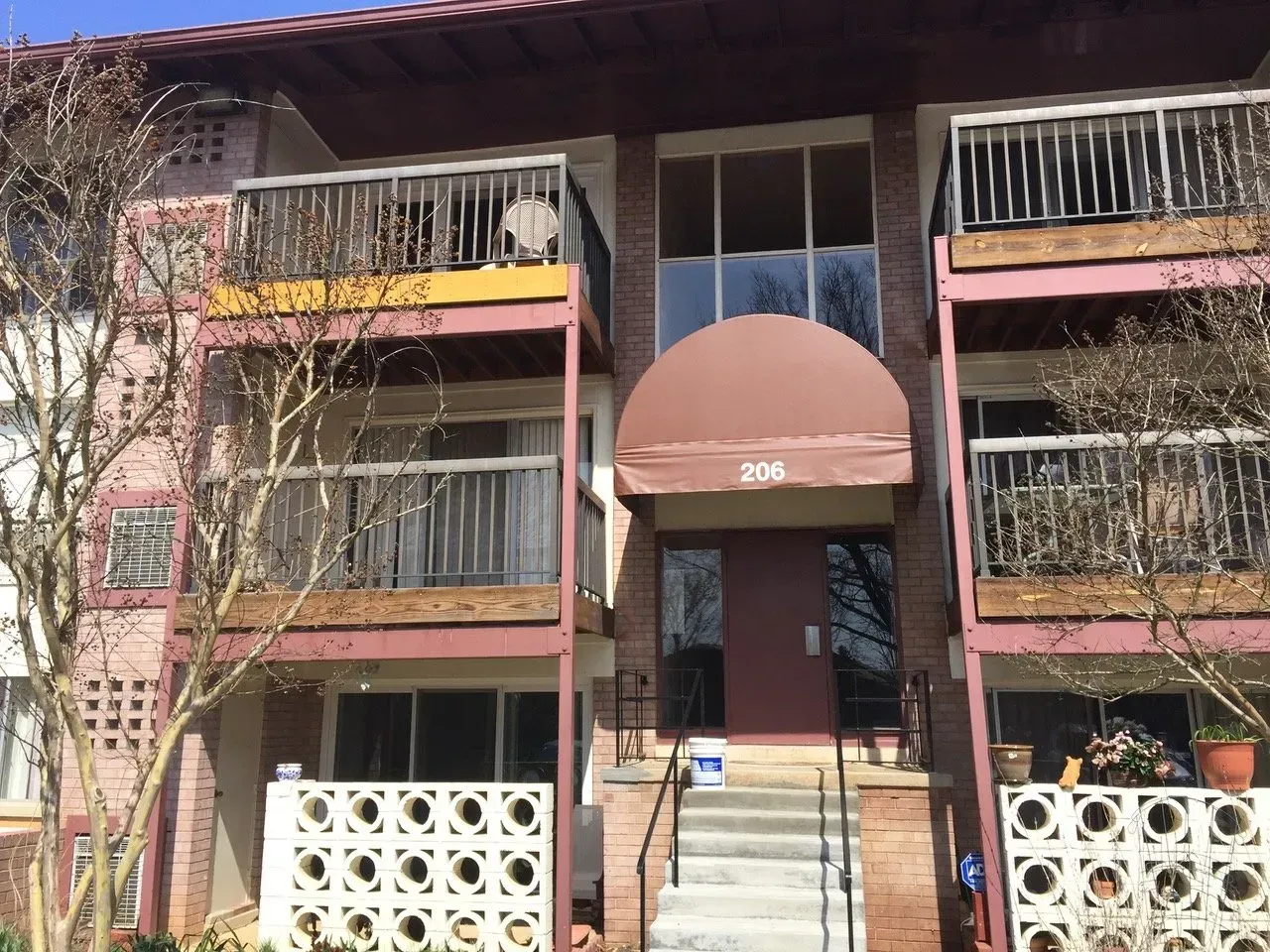 Three-story apartment building with a central staircase and balconies shaded by a brown awning.