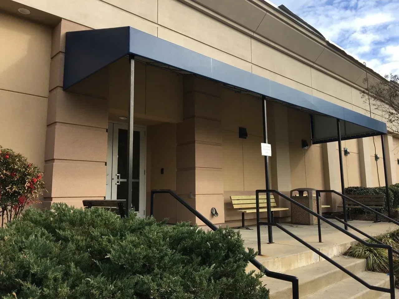 Building entrance with blue awning, ramp, steps, and benches beside shrubs